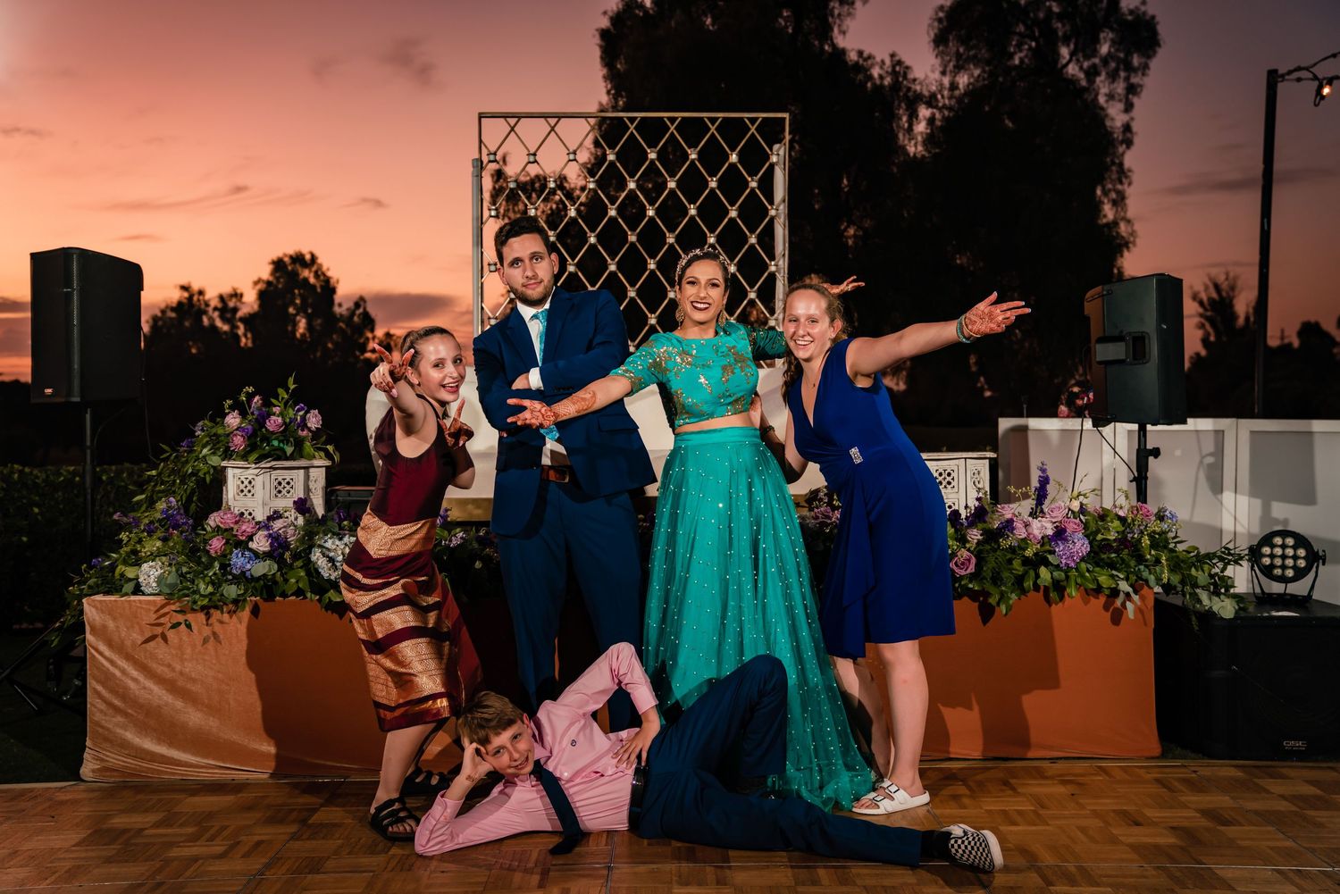 A group in formal attire strikes playful poses on a dance floor at sunset with decorative planters in the background.