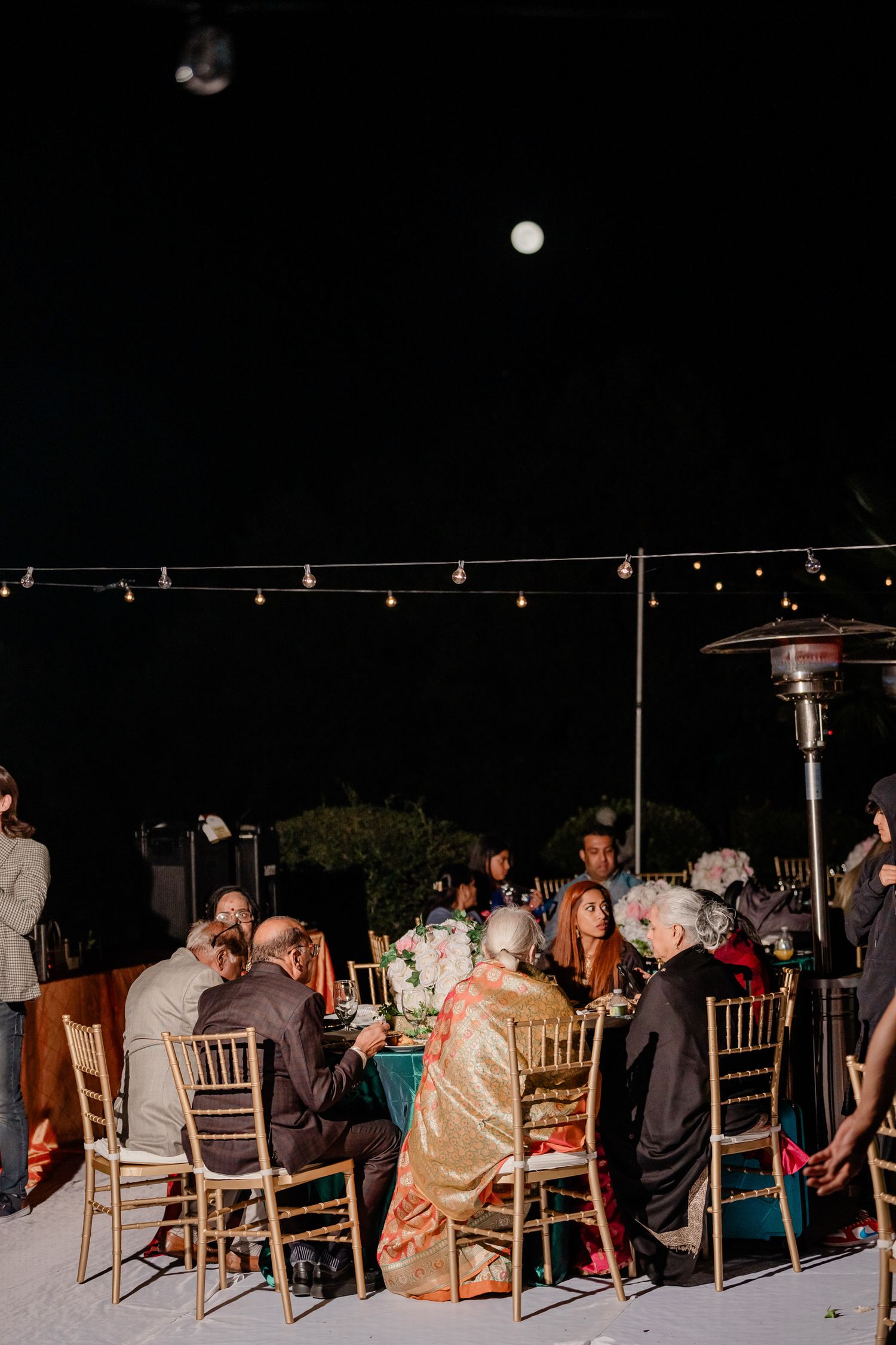 Wedding guests seated at outdoor reception tables with string lights overhead at night.