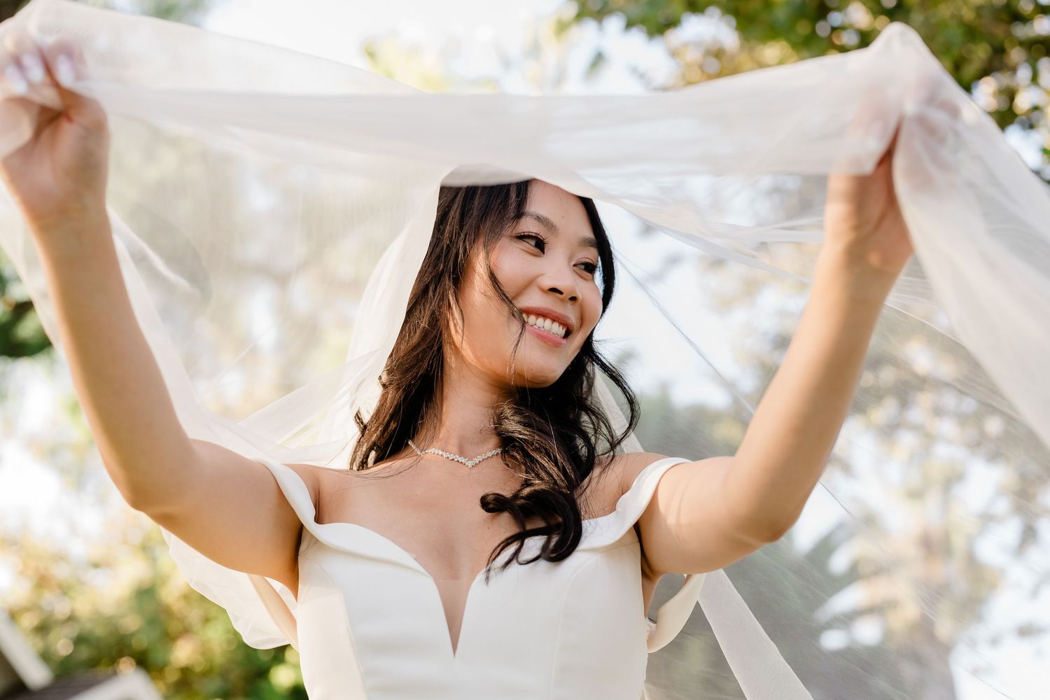 Bride joyfully holding up her wedding veil in a sunny outdoor setting.