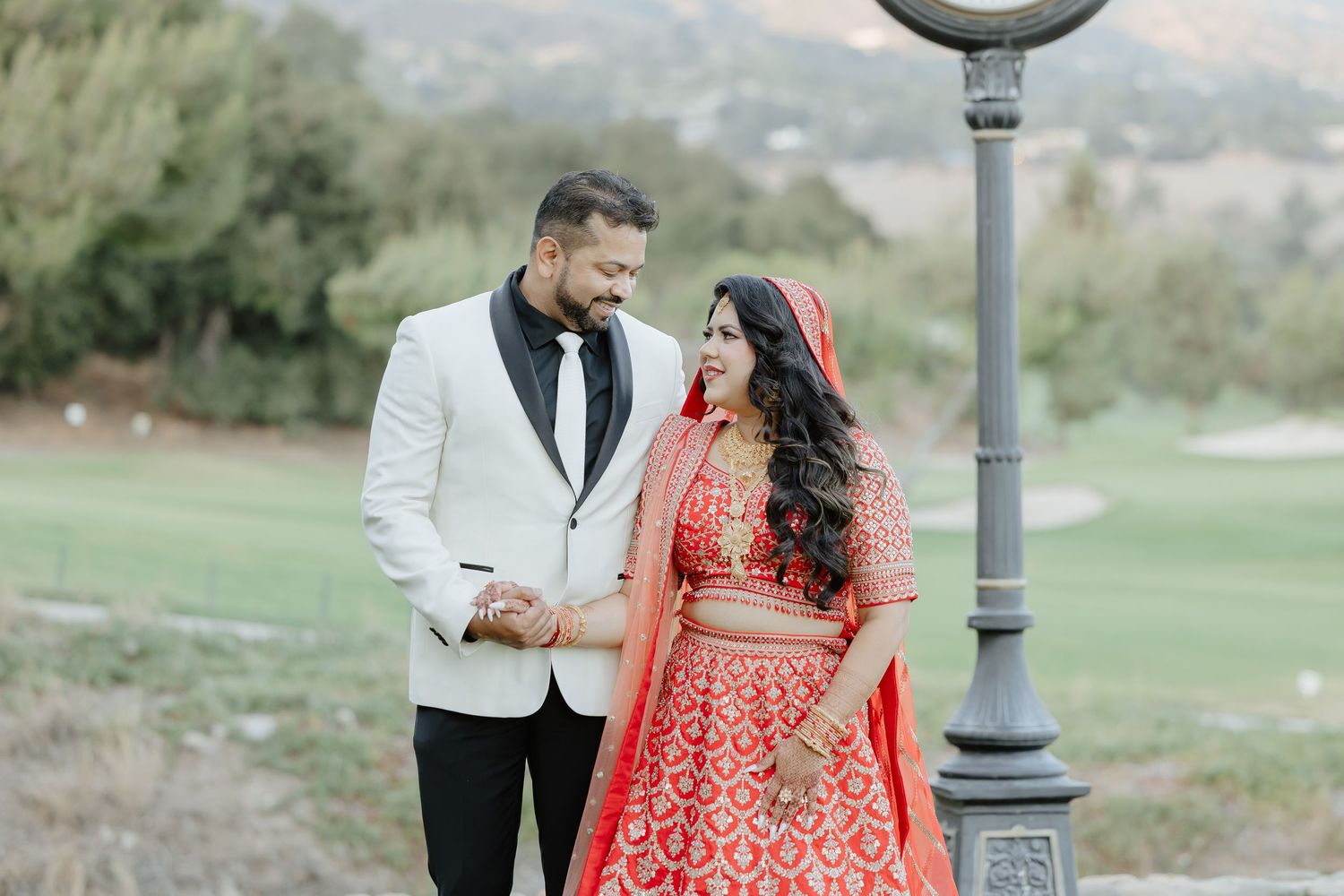 Couple in traditional Indian wedding attire, man in white tuxedo, woman in red lehenga, holding hands outdoors.