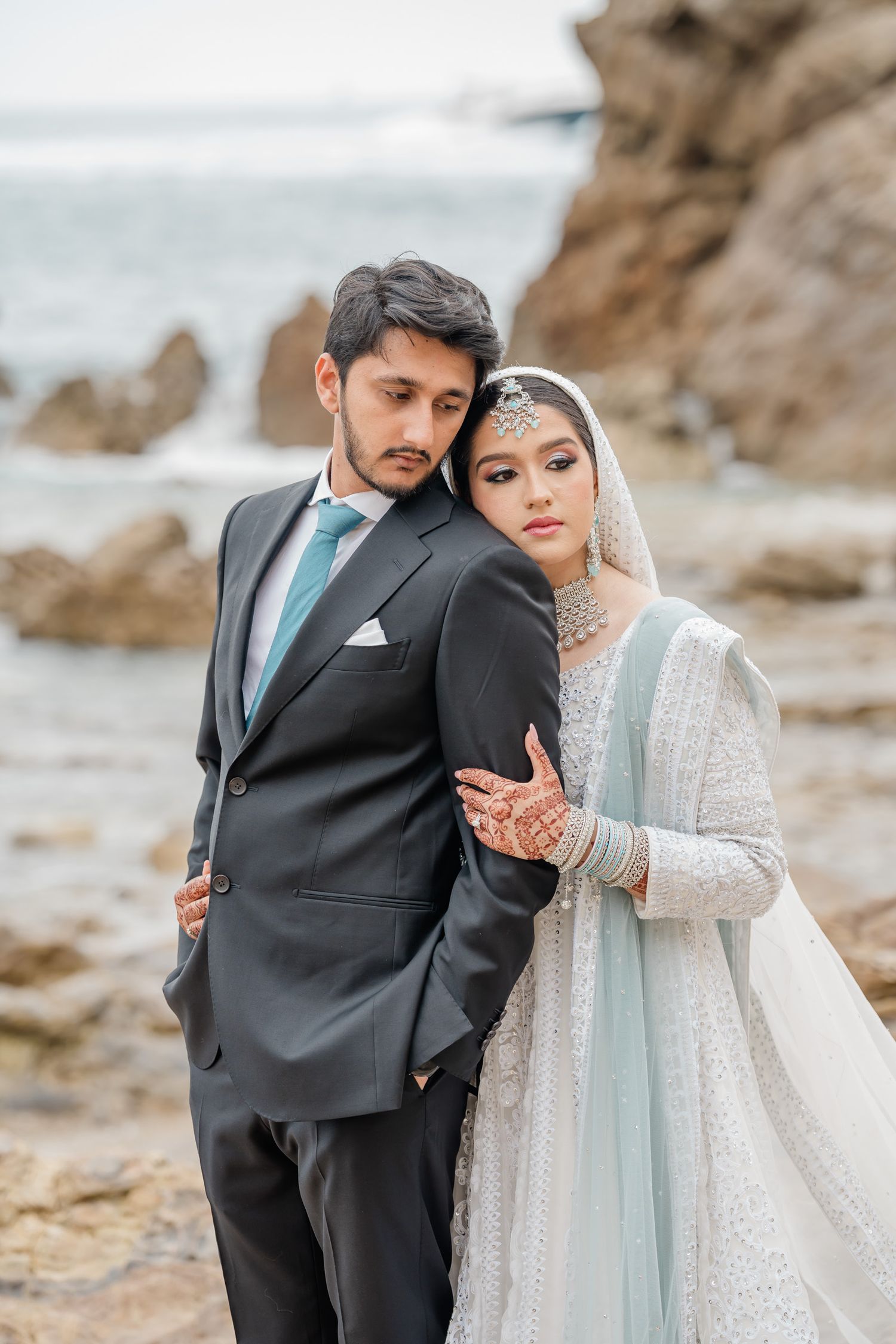 A couple in wedding attire poses together on a rocky beach, with the bride wearing a light blue and white traditional dress.