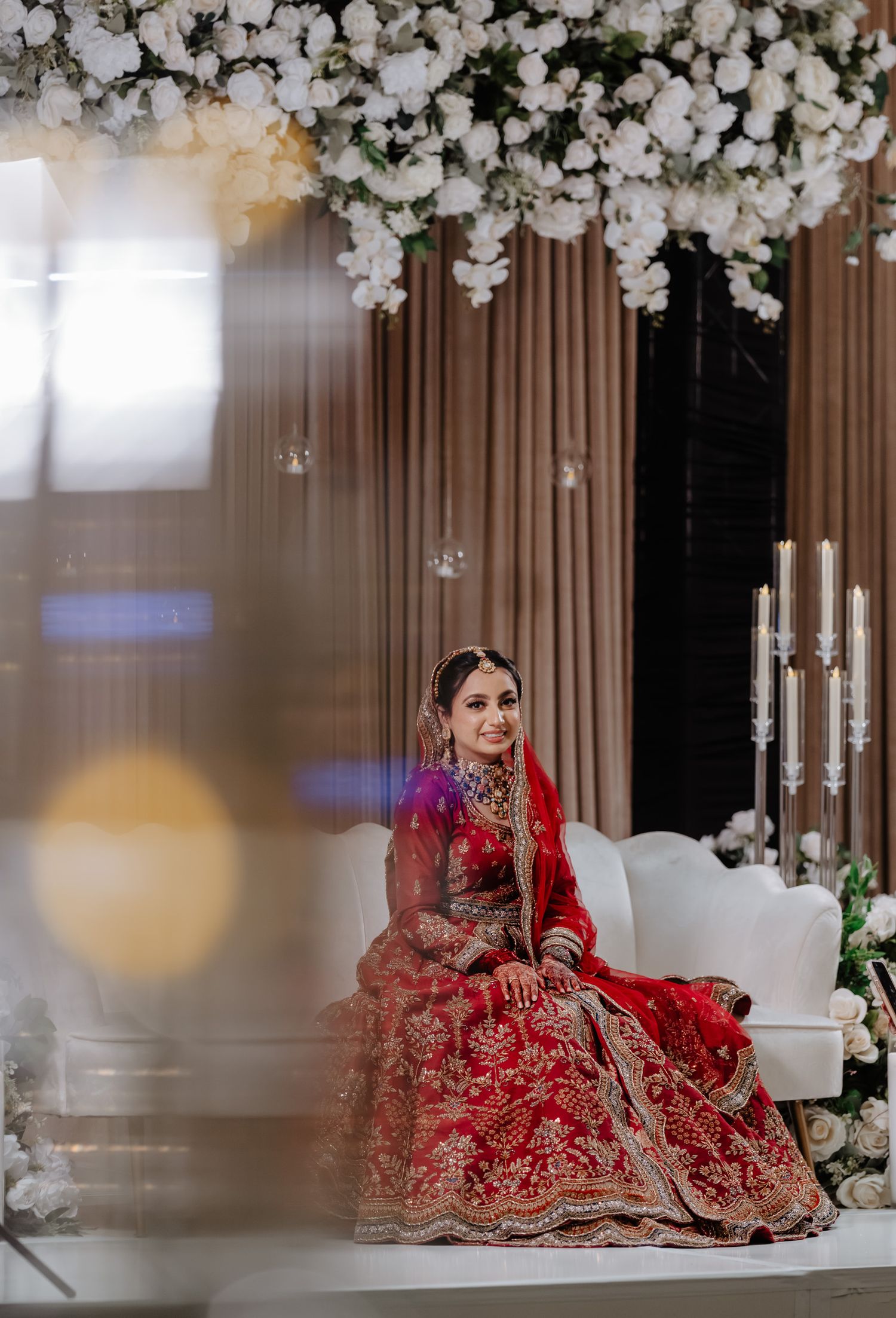Traditional Indian bride in red lehenga poses under elegant white floral arch and curtain backdrop.