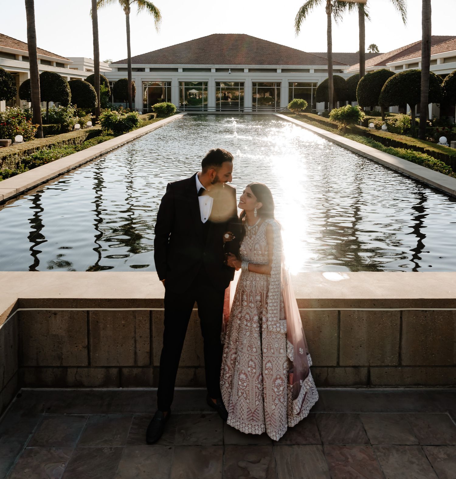 Couple shares a romantic moment by a reflecting pool surrounded by palm trees and architecture.