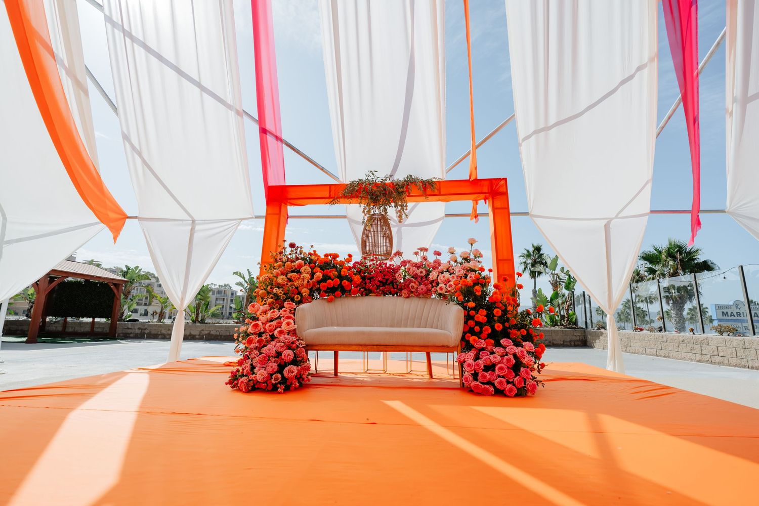 Vibrant Indian wedding mandap with orange floral arch, white sofa, and colorful draping at an outdoor venue.