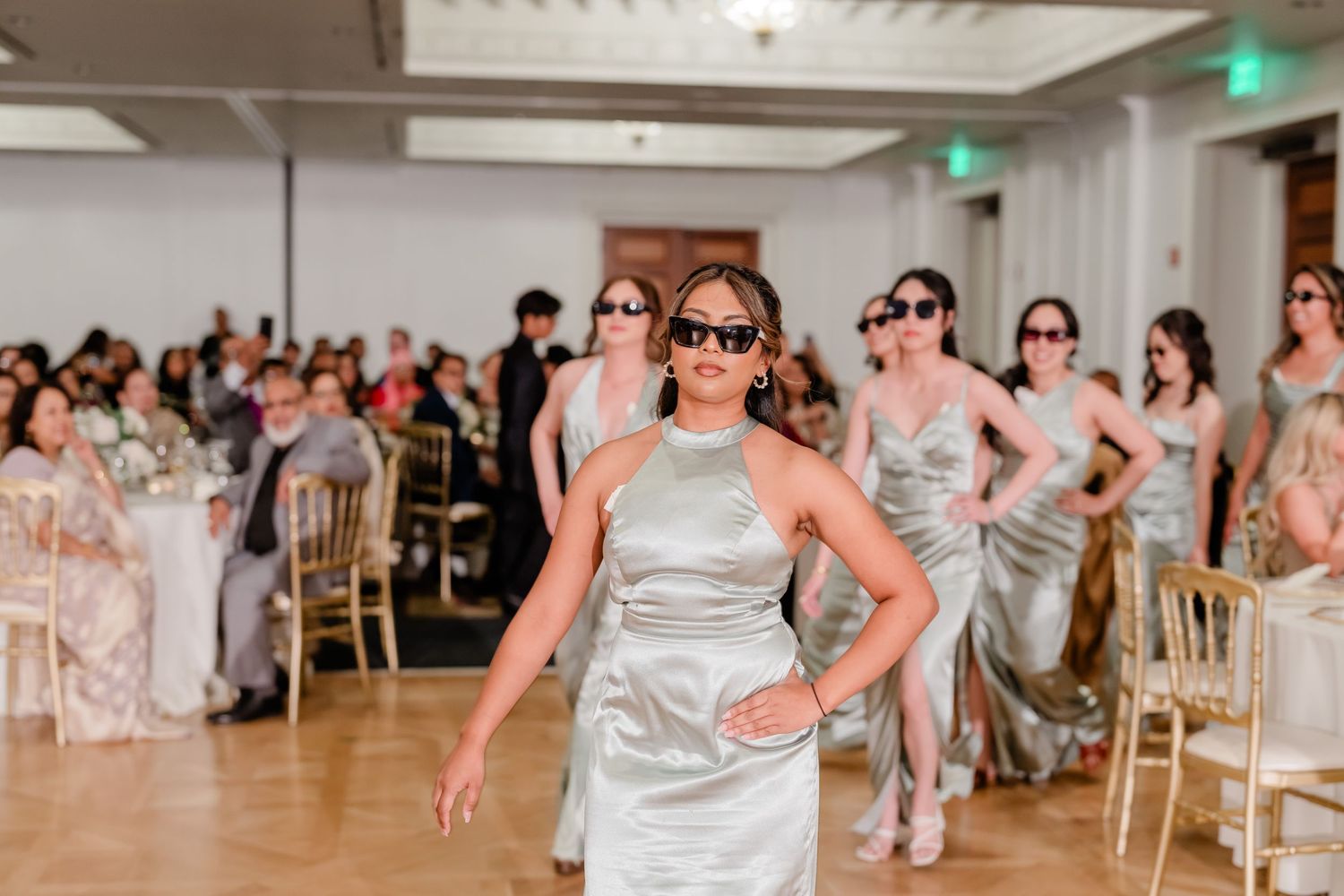 Wedding party members perform a synchronized dance routine in silver dresses on a ballroom floor.