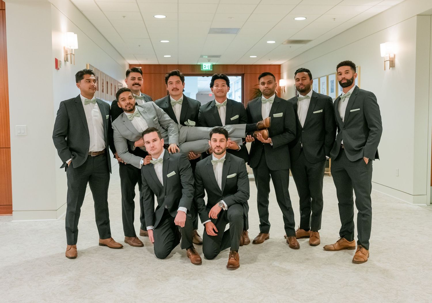 Groomsmen in matching dark suits and brown shoes pose together in a modern hallway.