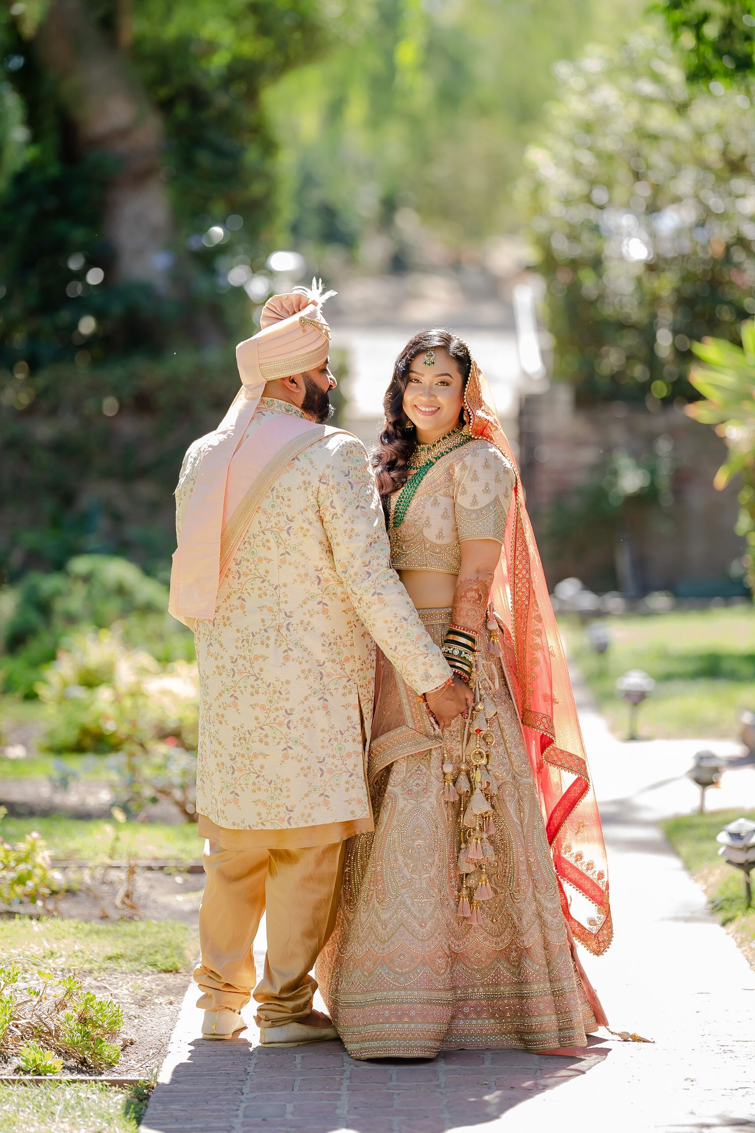 Couple in traditional Indian wedding attire walking together in a sunny garden setting.