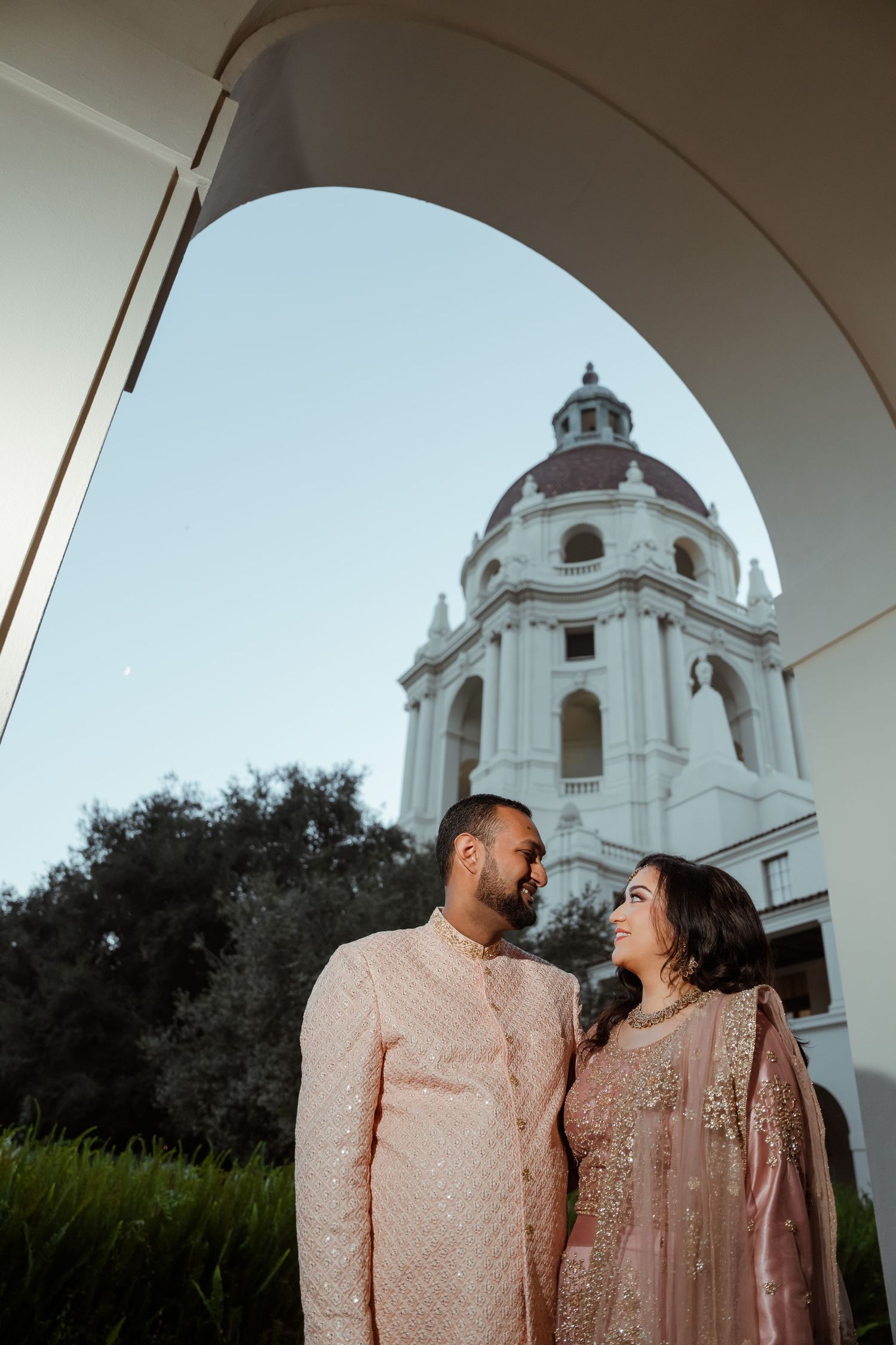 A couple in traditional South Asian attire pose beneath an archway of a white domed church building.