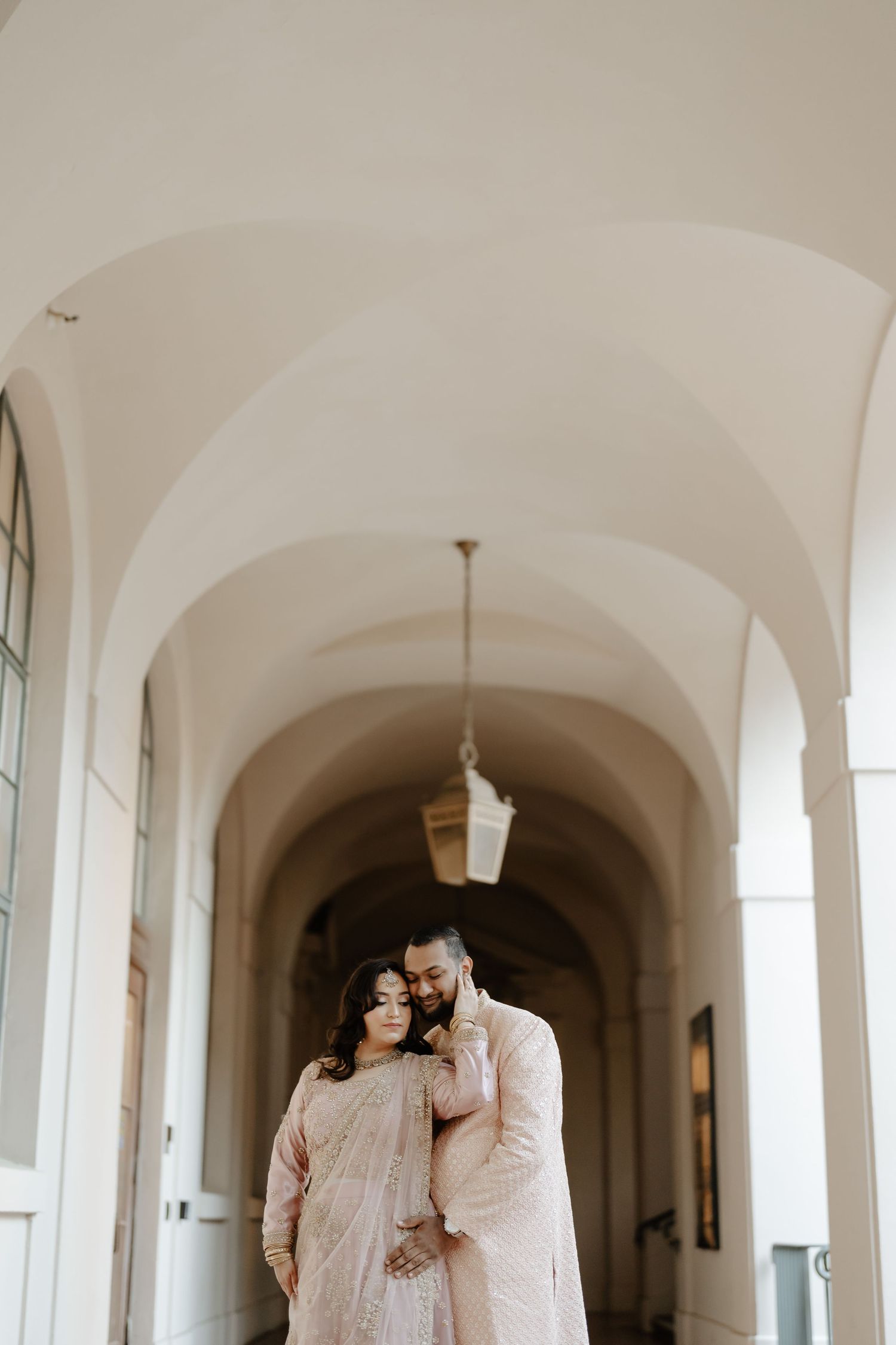 A couple in traditional cream outfits poses in a grand arched corridor.