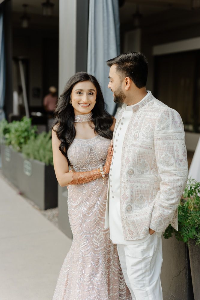 Indian couple in elegant wedding attire, bride in beaded gown with mehndi, groom in white embroidered sherwani.