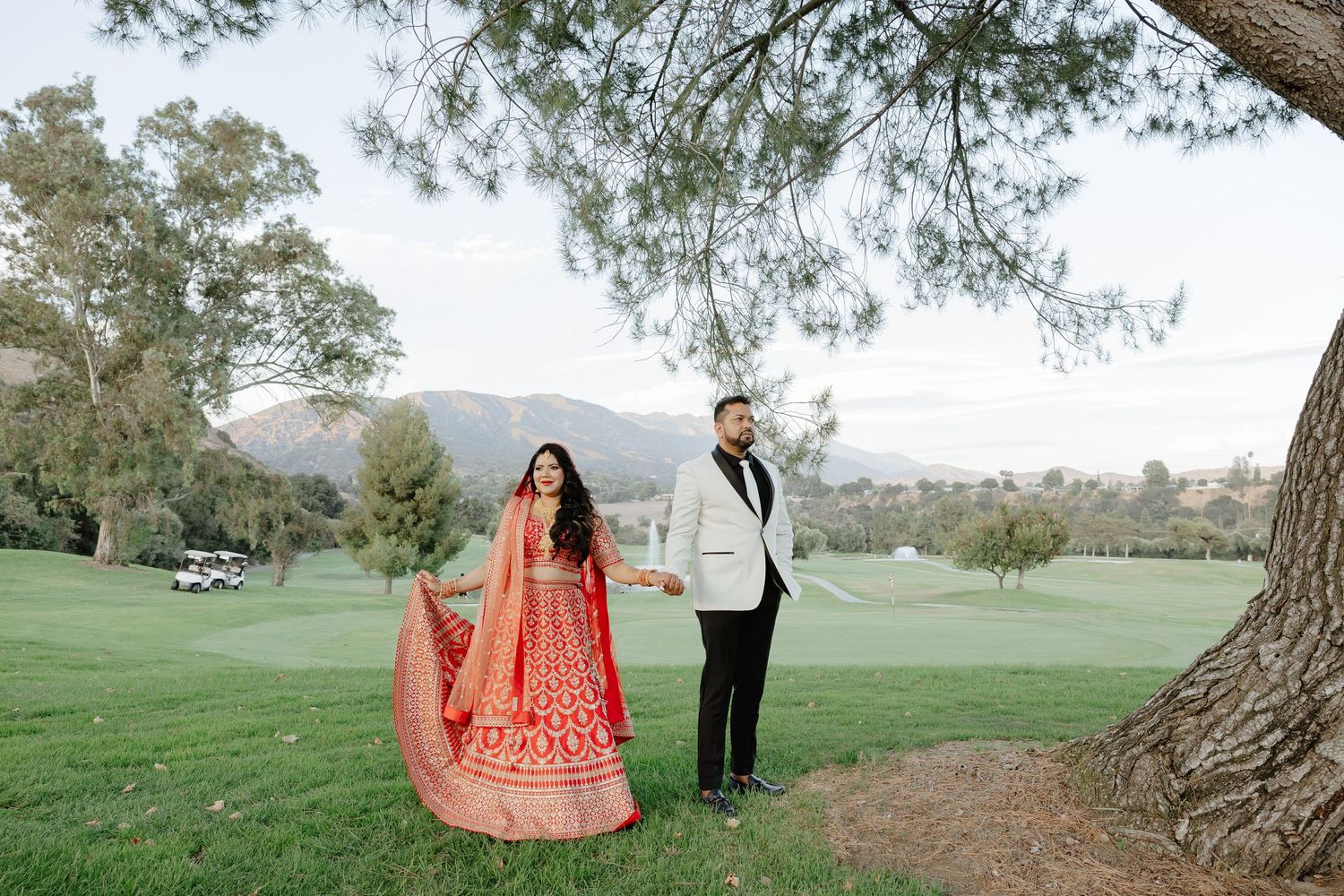 Couple in traditional Indian wedding attire holding hands on a lush golf course with mountain views.