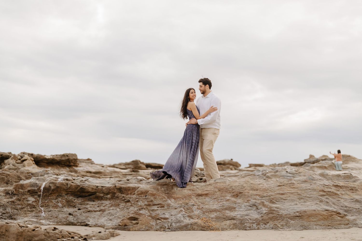 Couple embracing on rocky coastal cliffs with overcast sky, woman in flowing blue dress, romantic engagement photo.