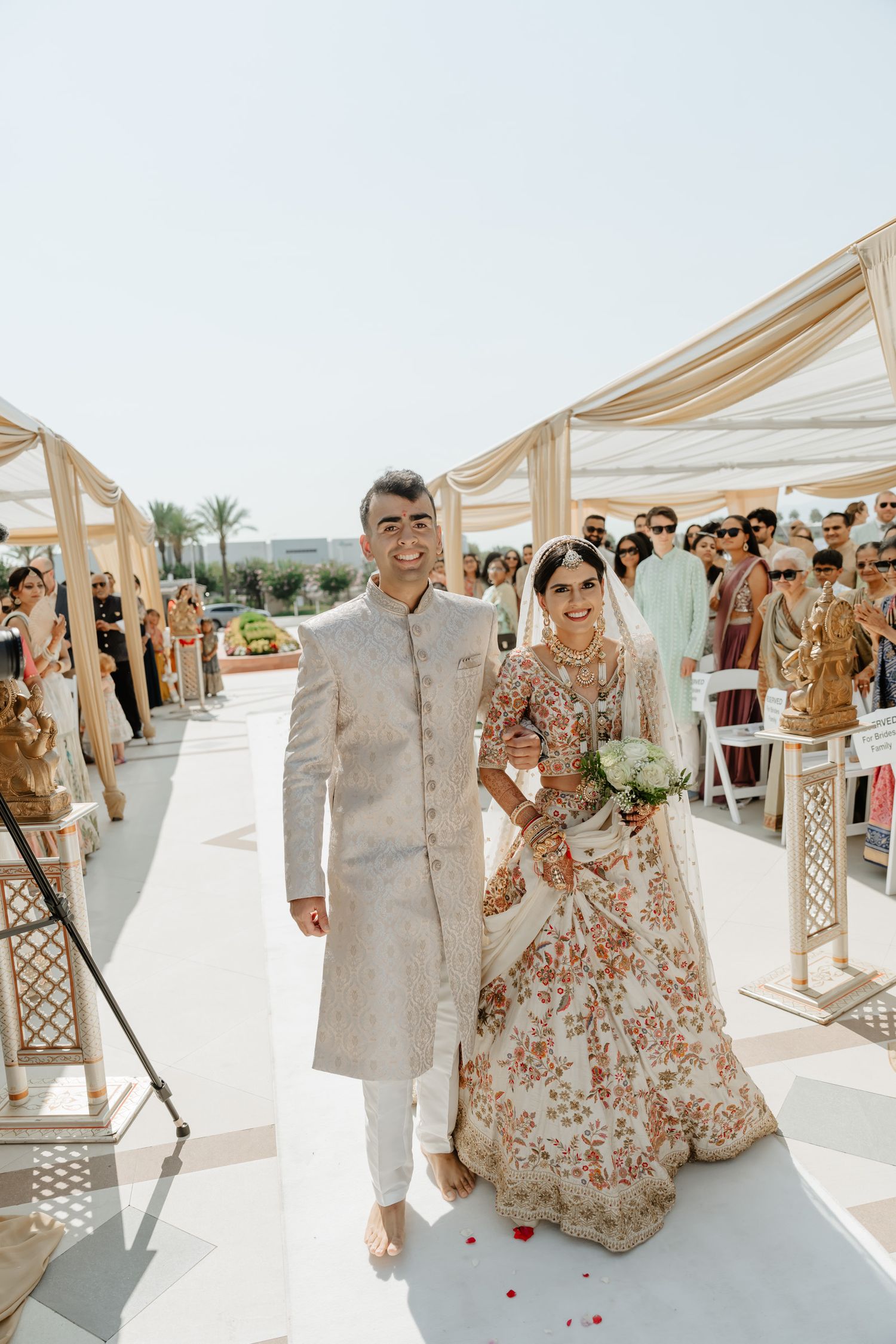 Wedding couple walks under beige canopy structures on sunny day with guests in background.