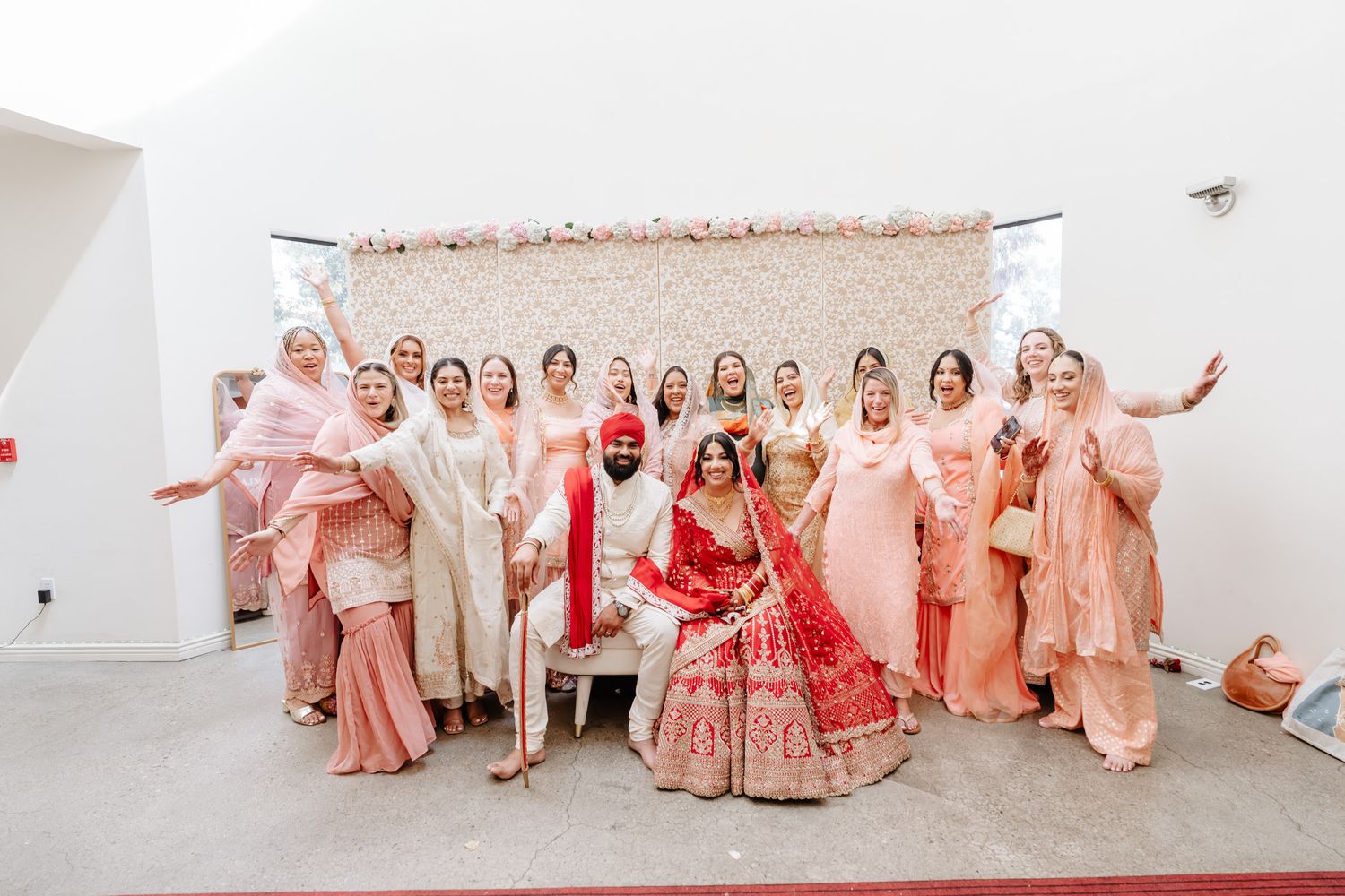 Wedding party group photo with bridesmaids wearing pink outfits against white backdrop.