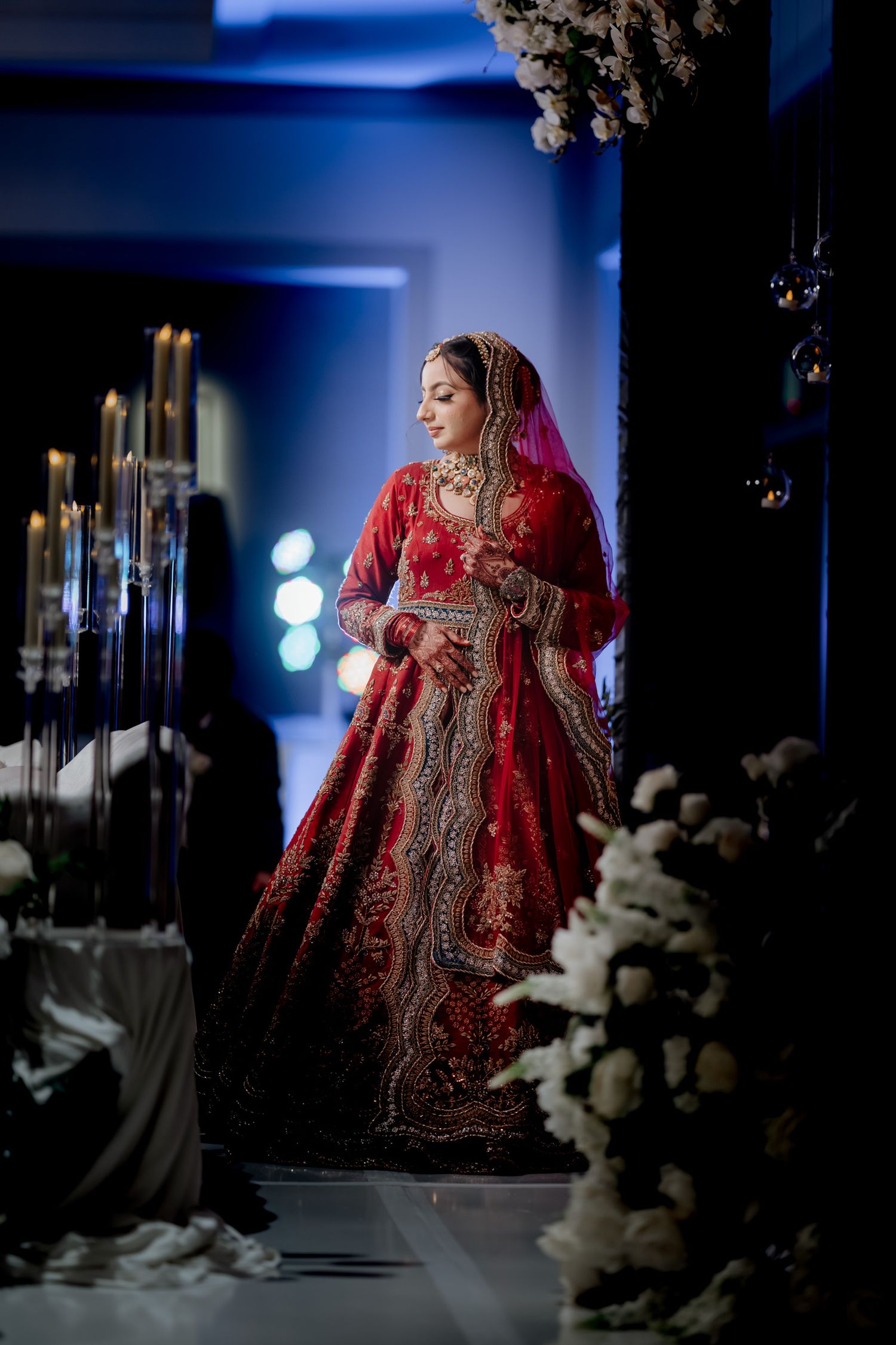 A bride in an ornate red lehenga poses dramatically in a blue-lit room with white floral decorations.