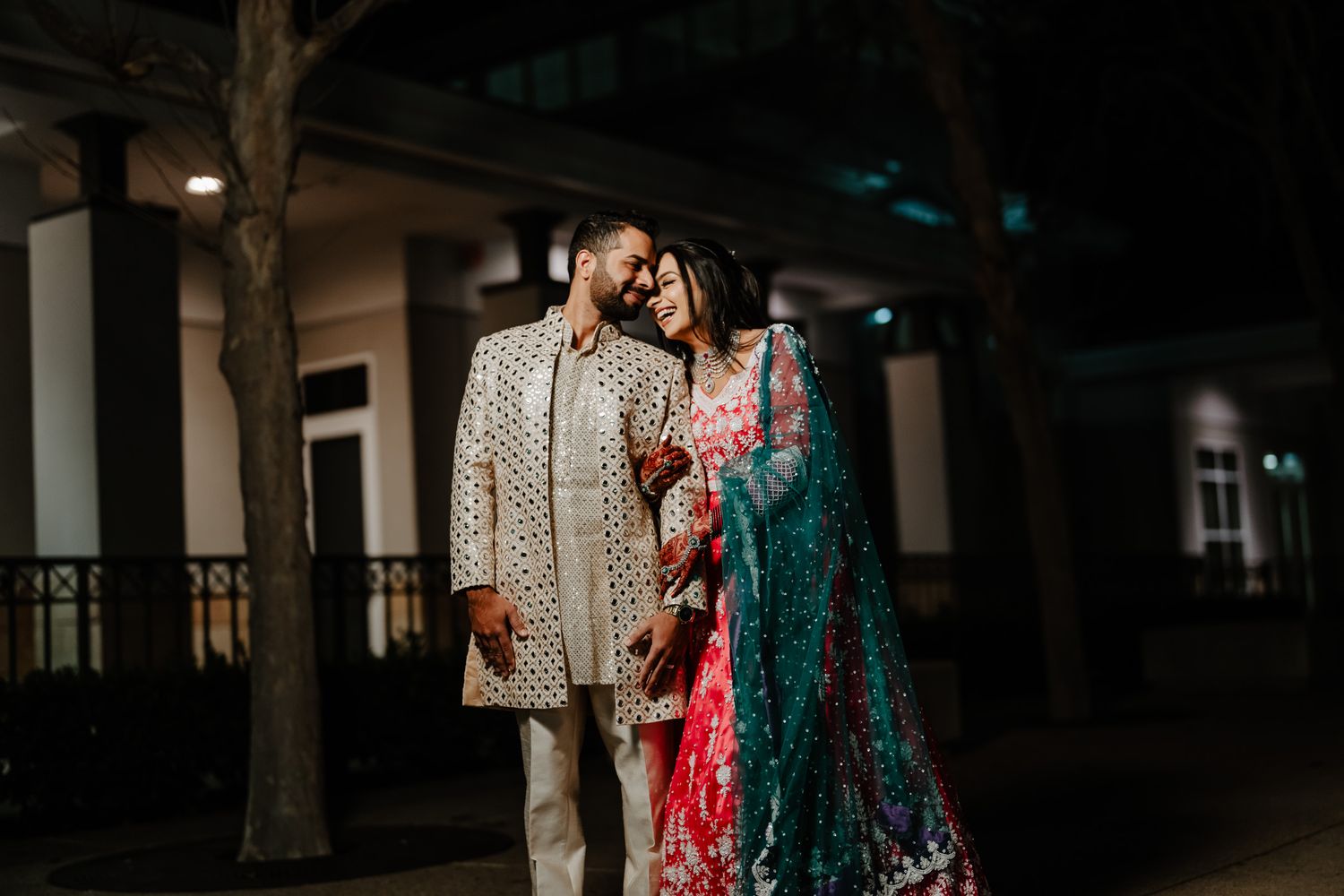 A couple in traditional wedding attire poses together at night under outdoor lighting.