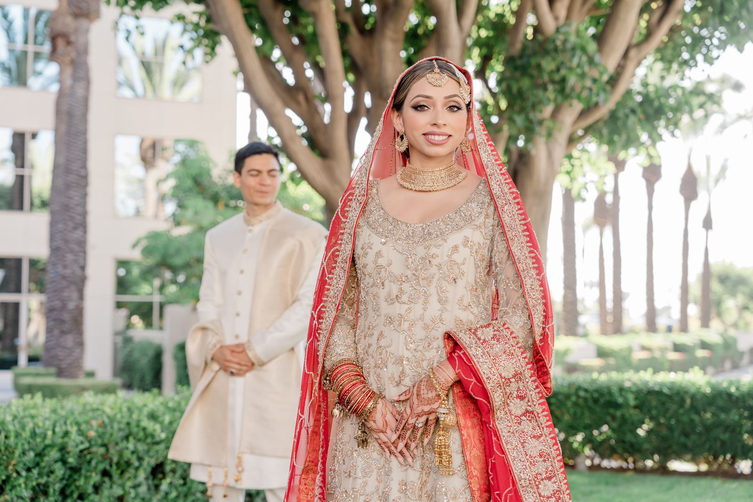 Traditional South Asian bride in embellished cream and red wedding attire poses outdoors with groom in background.