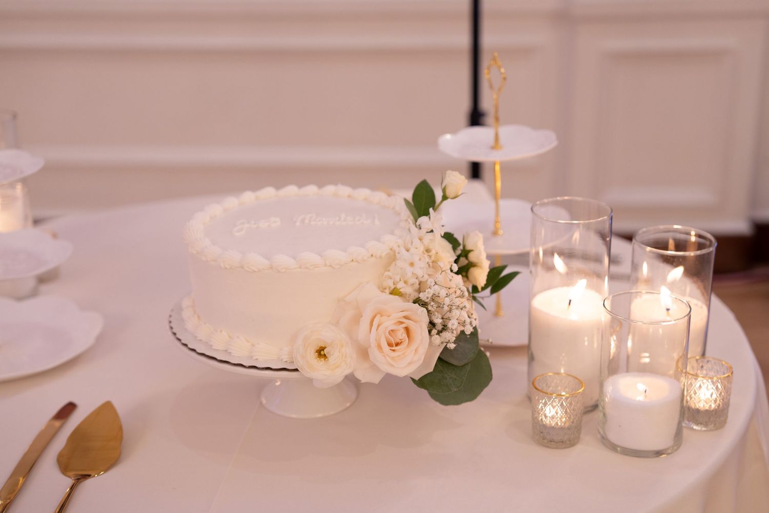 A white wedding cake decorated with roses sits beside glowing candles on an elegant table setting.