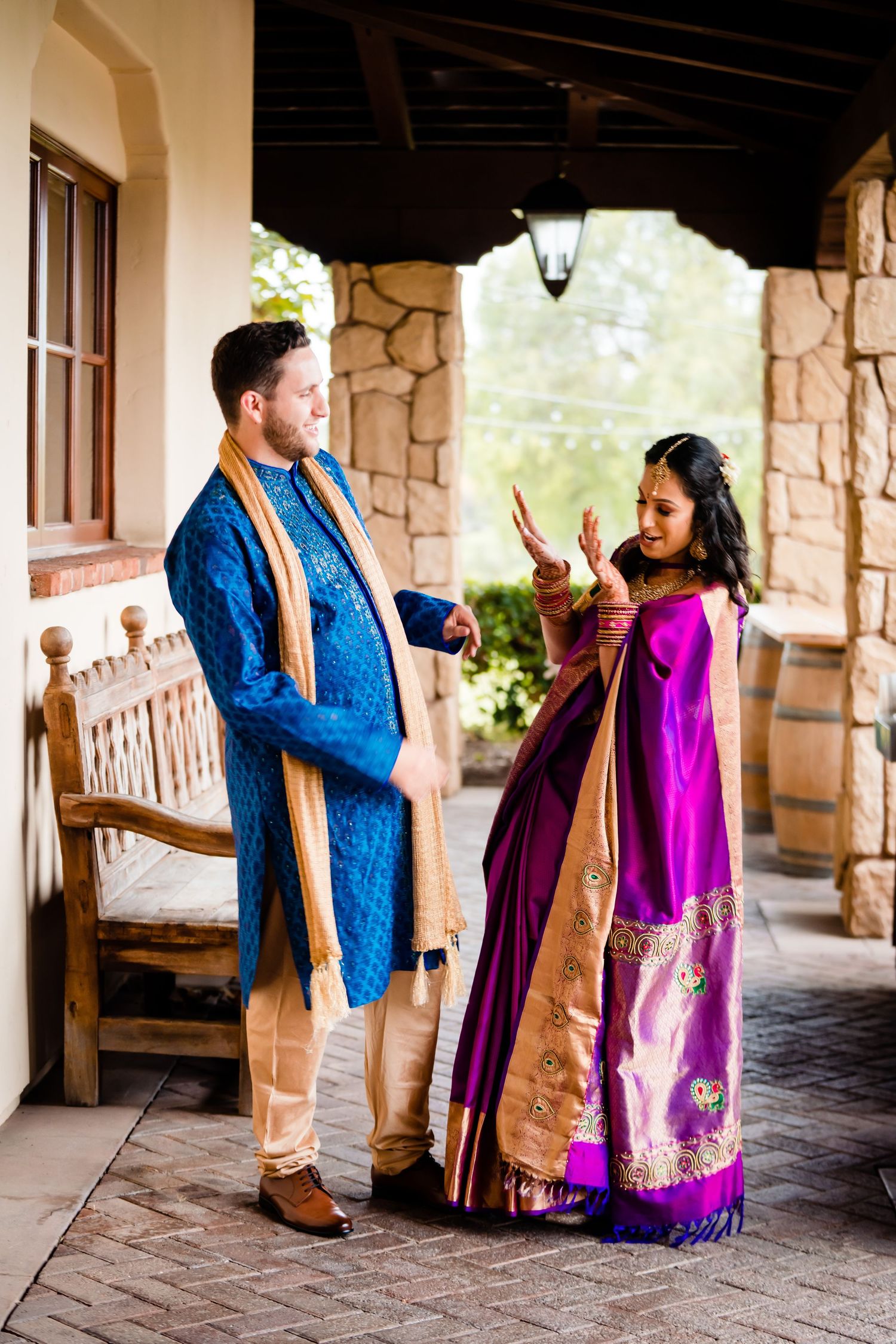 A couple in traditional Indian and Western fusion attire share joyful moments in a stone-columned outdoor walkway.