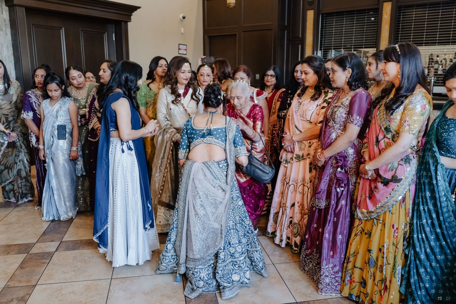 Women in colorful traditional Indian attire gather indoors, with a bride in silver lehenga at the center of the celebration.