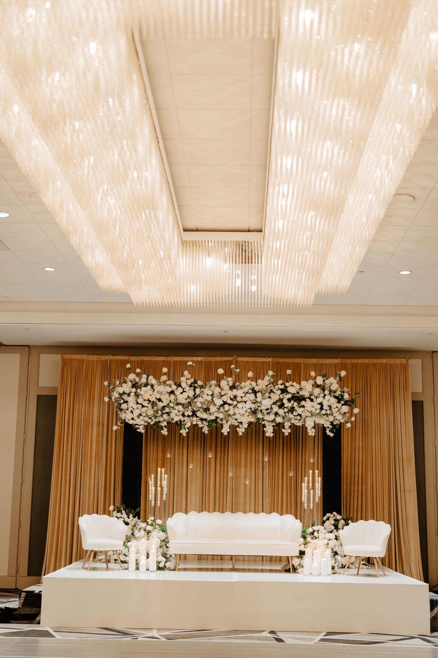 Modern wedding venue with crystal chandeliers and white floral arch backdrop for ceremony setup.