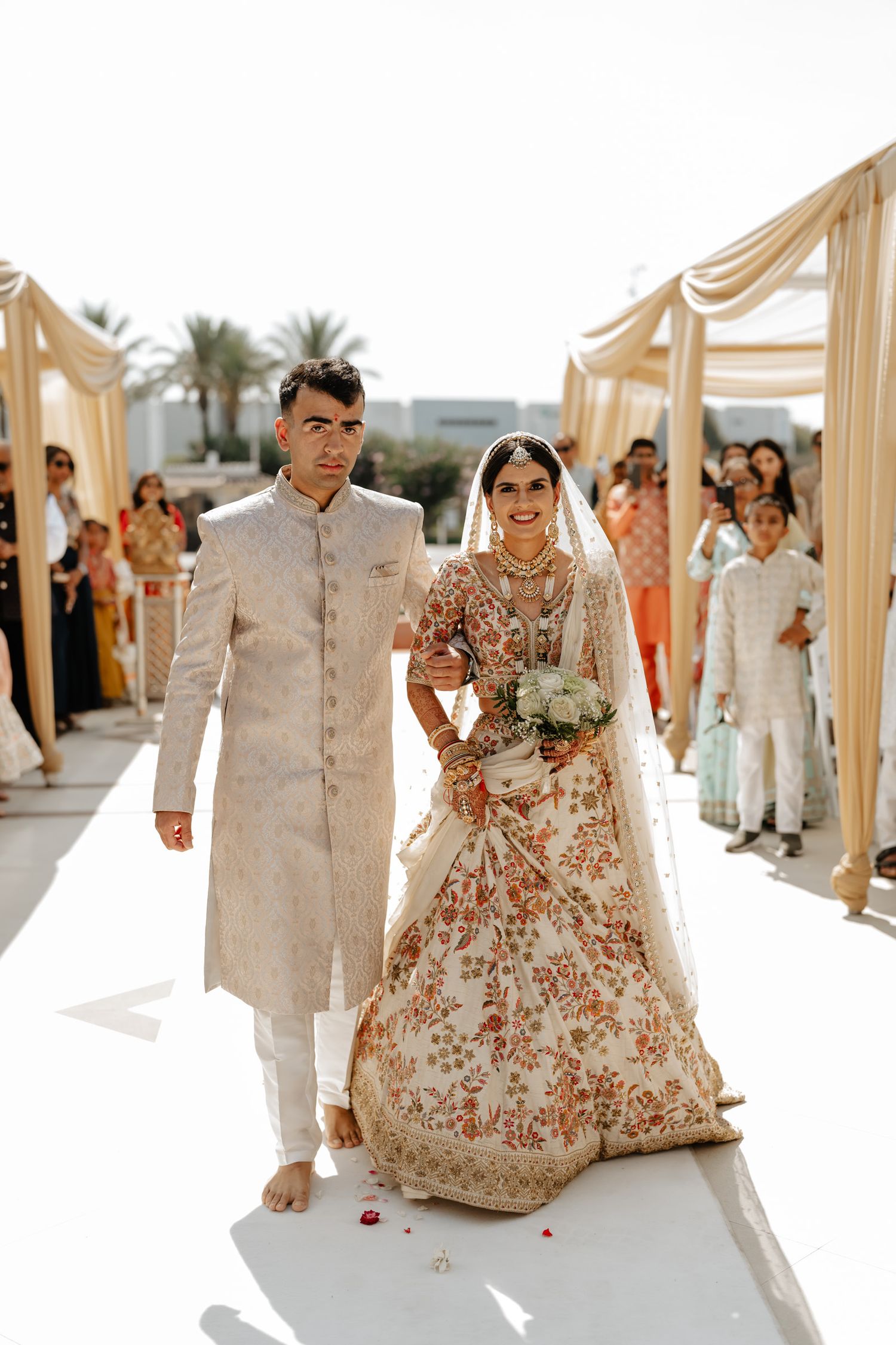 Traditional Indian wedding ceremony with couple in ornate attire walking down aisle under draped cream fabric canopy.
