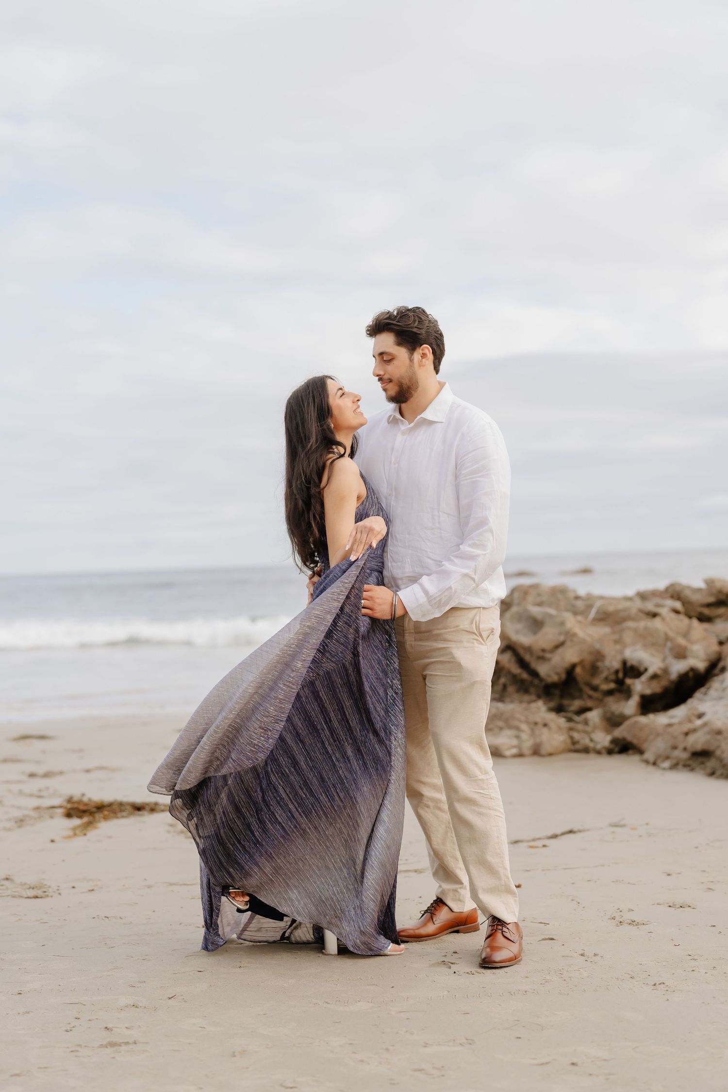 Couple embracing on a sandy beach, woman in flowing blue dress, man in white shirt and khaki pants, ocean backdrop.