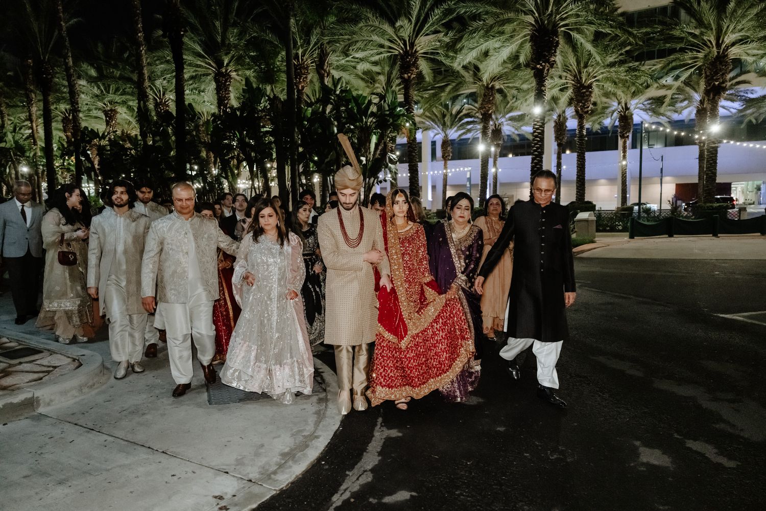 Wedding party in traditional South Asian attire standing together under palm trees at dusk.