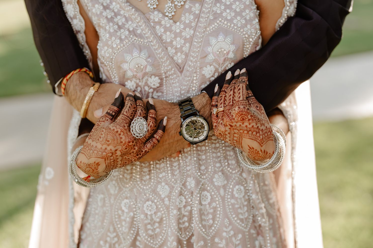 Elegant wedding couple holds hands showing off intricate henna designs and rings in natural outdoor setting.