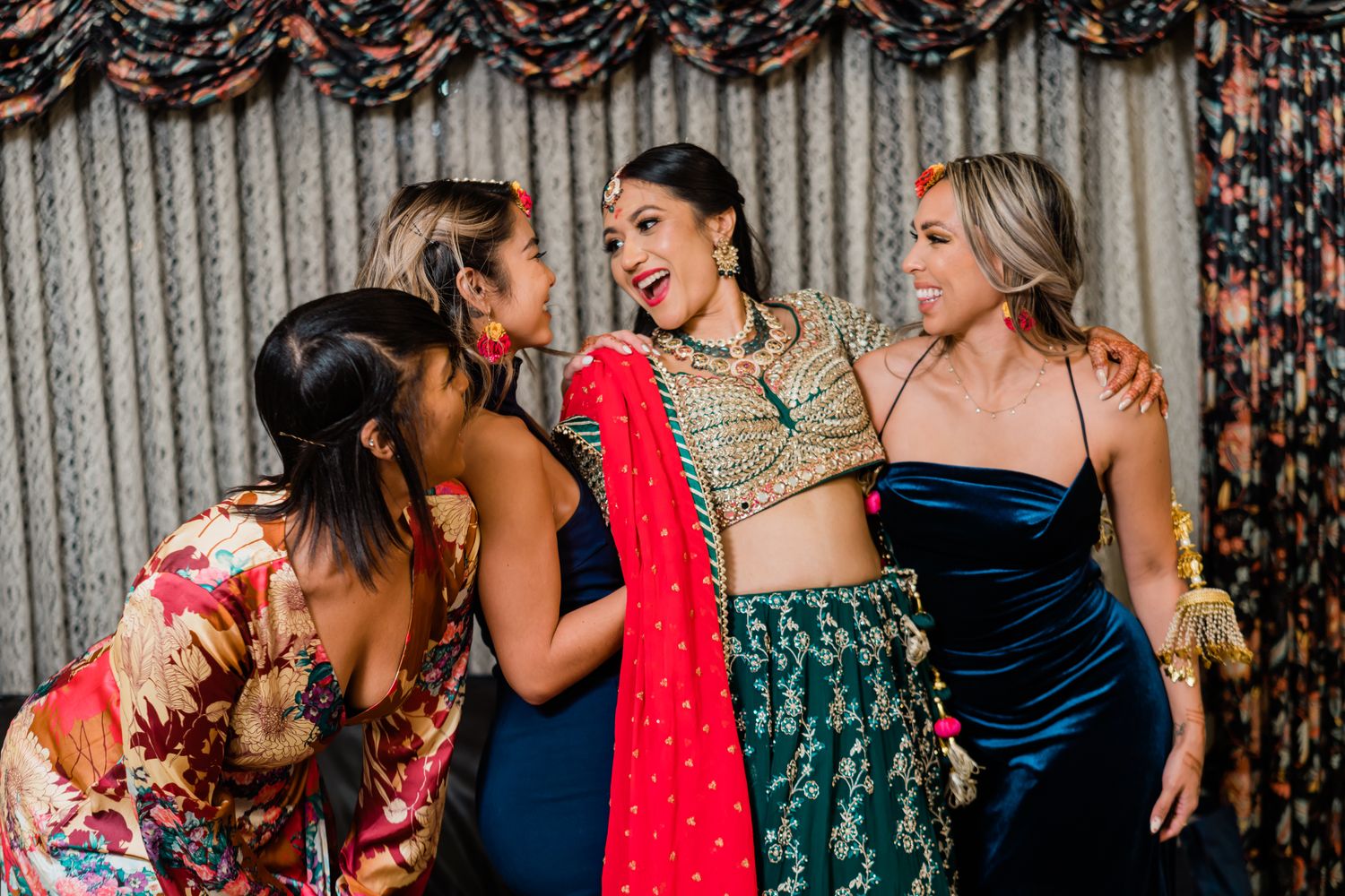 A group of women in formal dresses laugh and dance together at an elegant party with decorative curtains in the background.