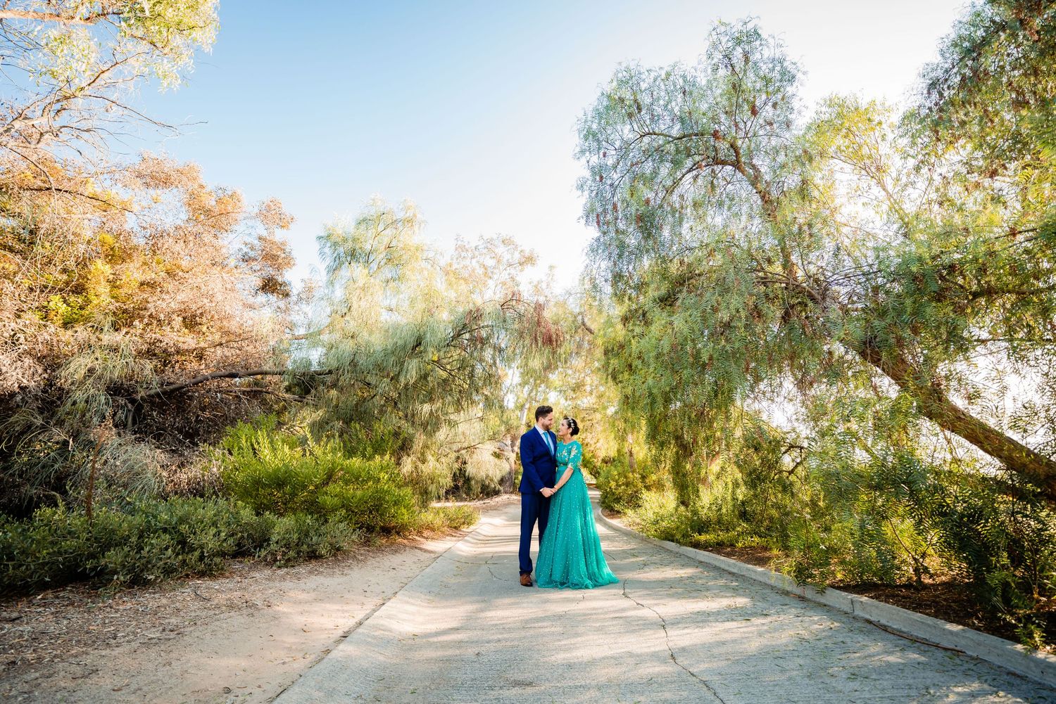 Couple in elegant formal wear walk along a tree-lined path during golden hour at a park.