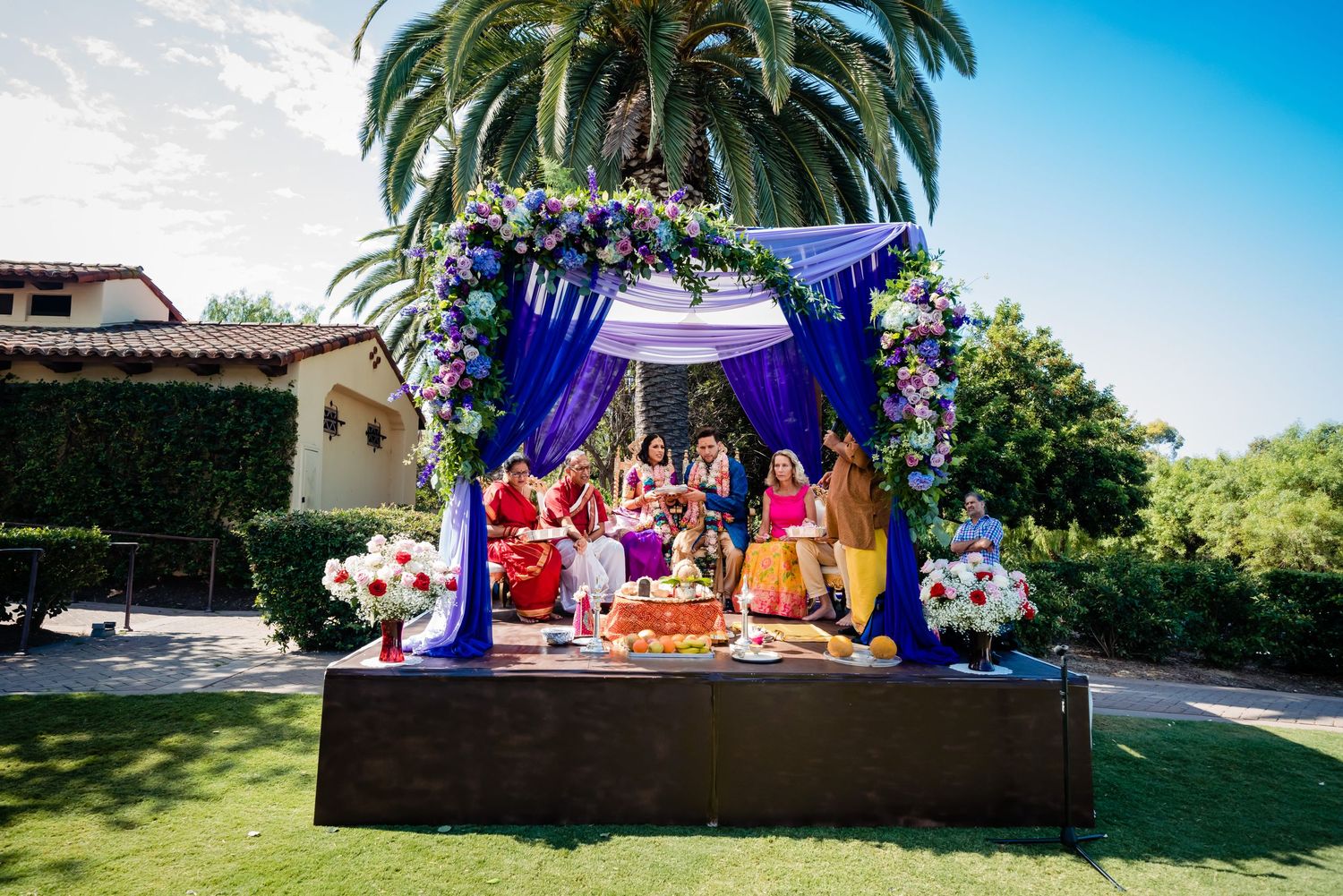 Colorful outdoor Indian wedding ceremony setup with purple mandap and palm trees in a garden setting.