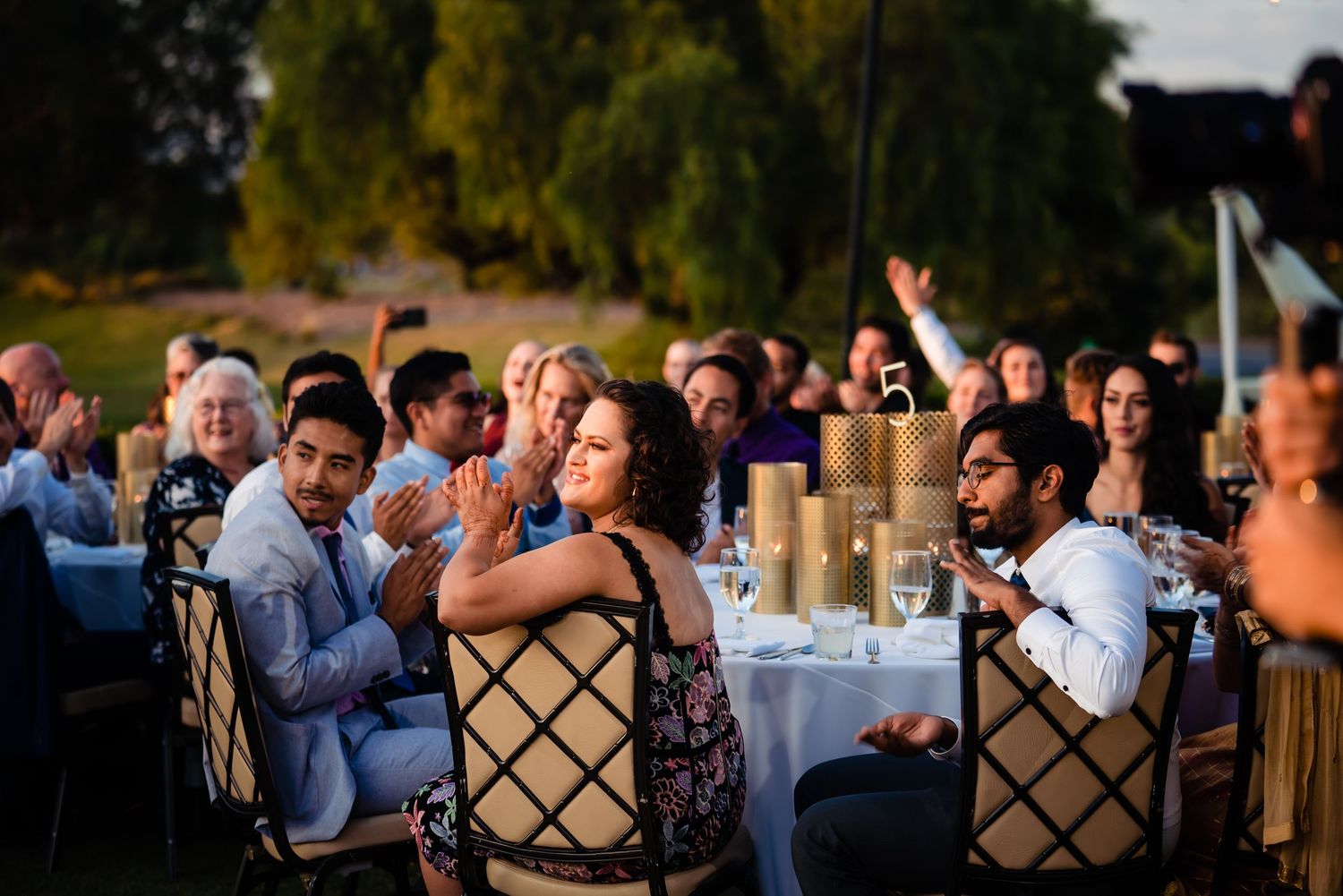 Wedding guests enjoy an outdoor reception celebration at sunset with tables and chairs set up in a garden setting.