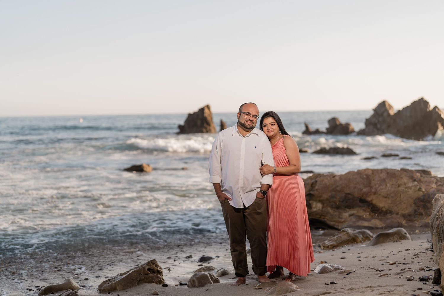 Couple walking along rocky beach at sunset with waves crashing against the shore in the background.