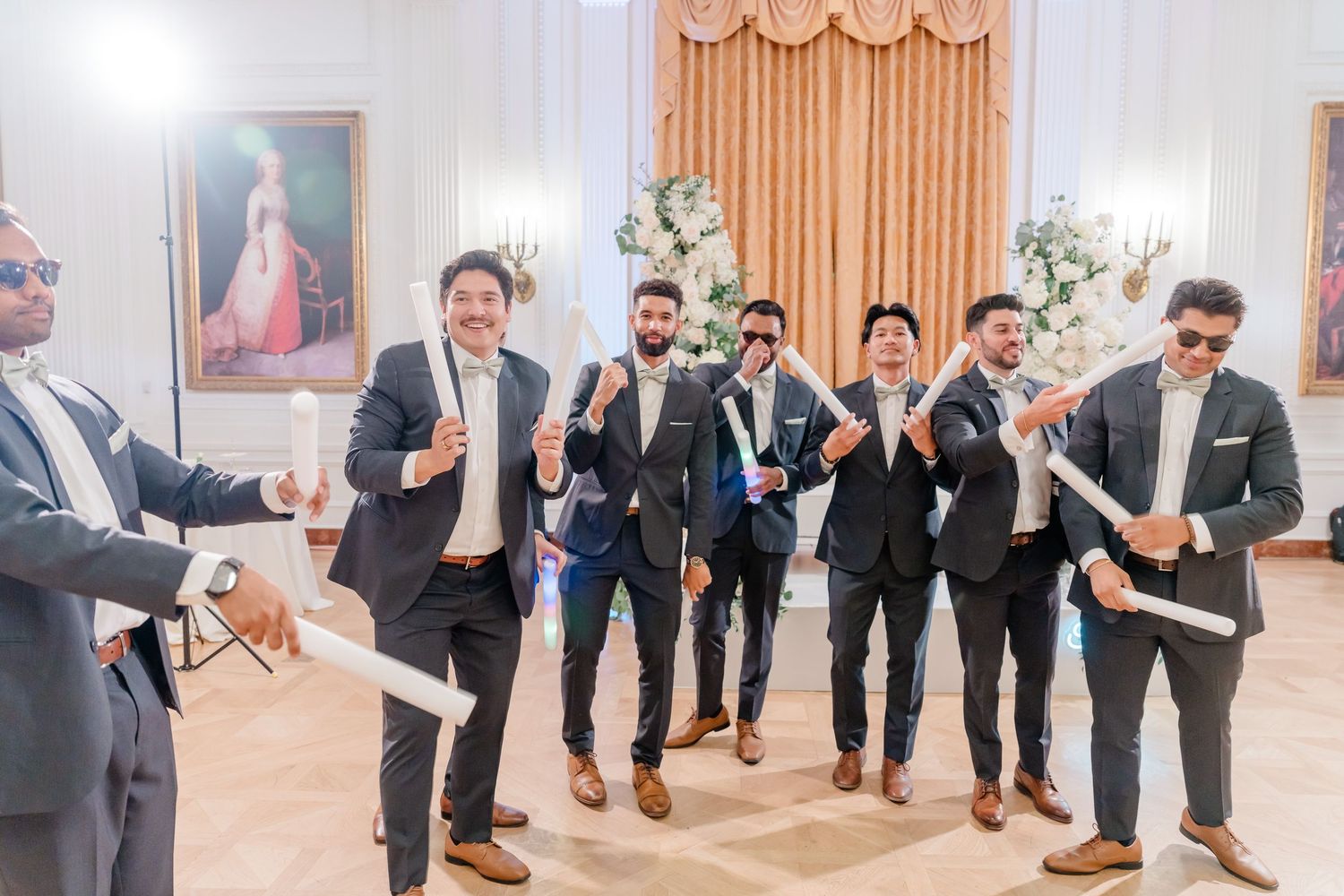 Wedding party groomsmen in gray suits performing synchronized dance moves in an elegant ballroom.