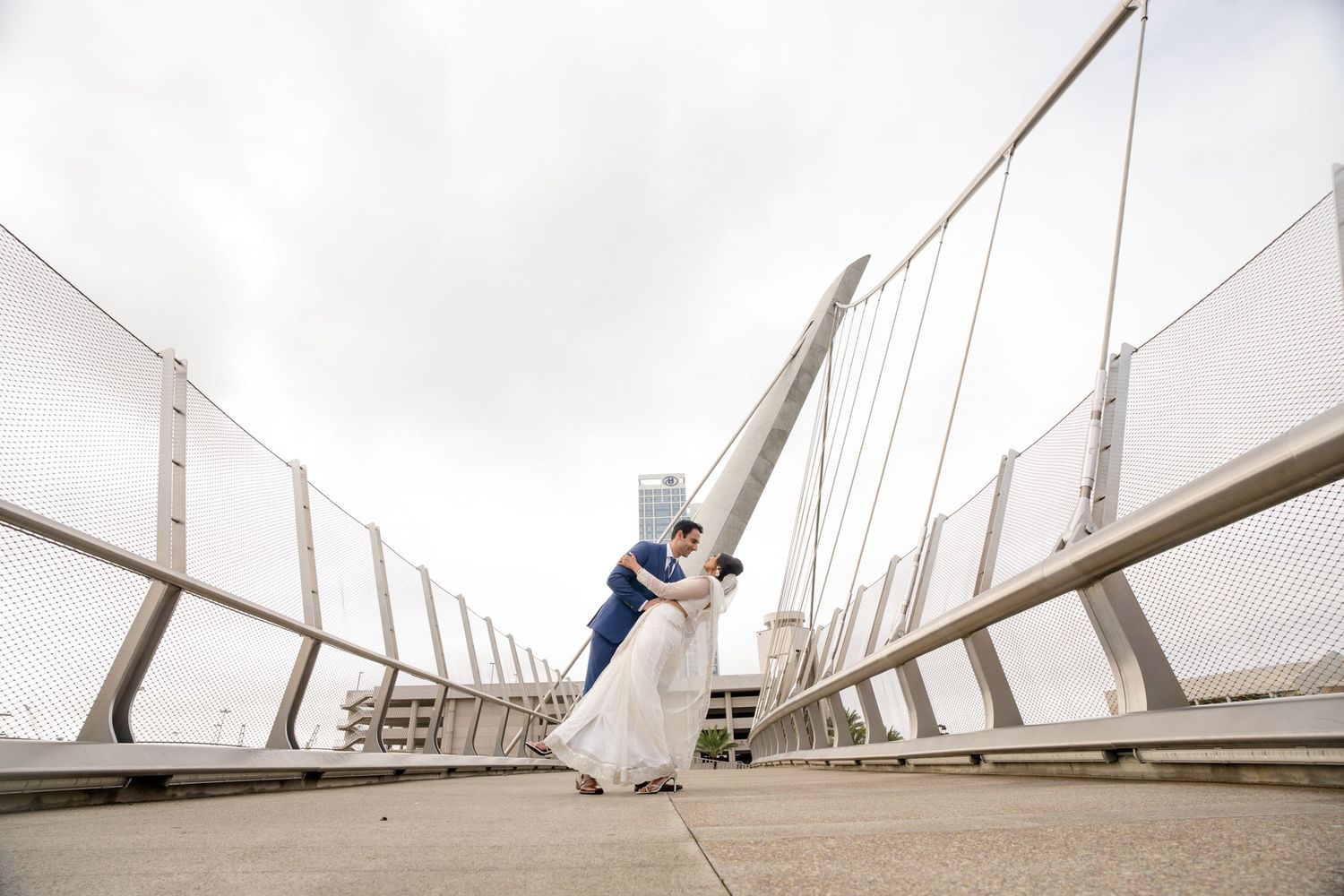 Bride and groom share a romantic dip kiss on a modern suspension bridge under an overcast sky.