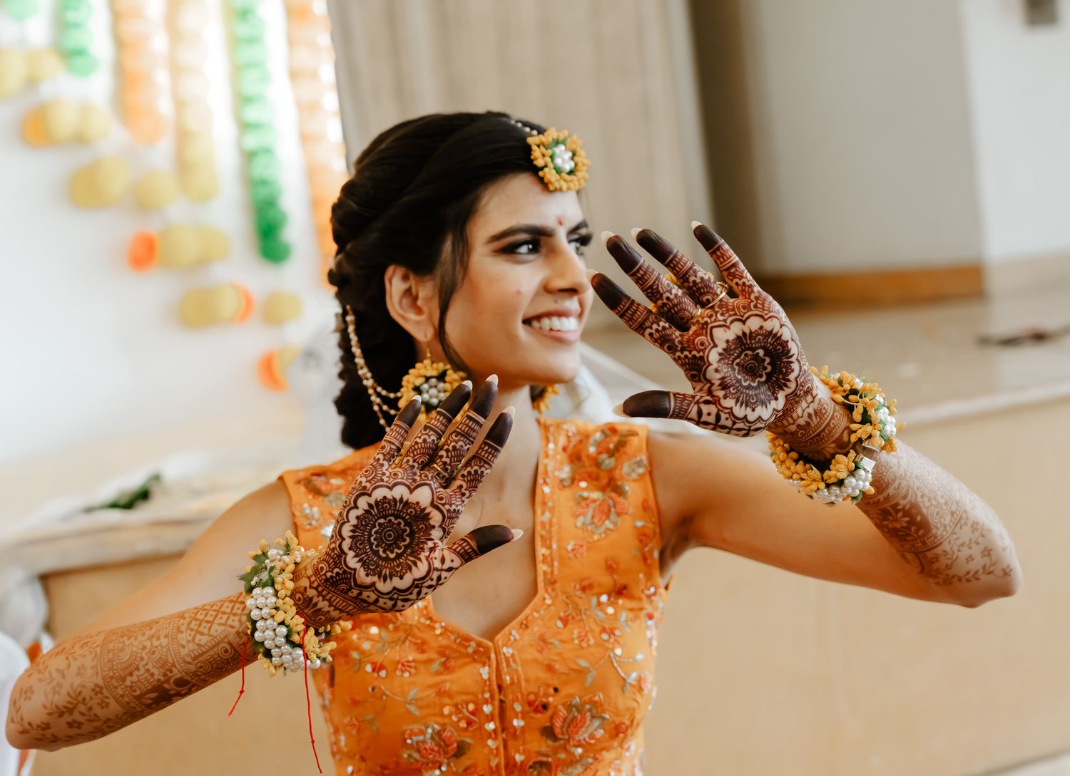 A person wearing an orange traditional outfit shows off intricate henna designs on their hands during a celebration.