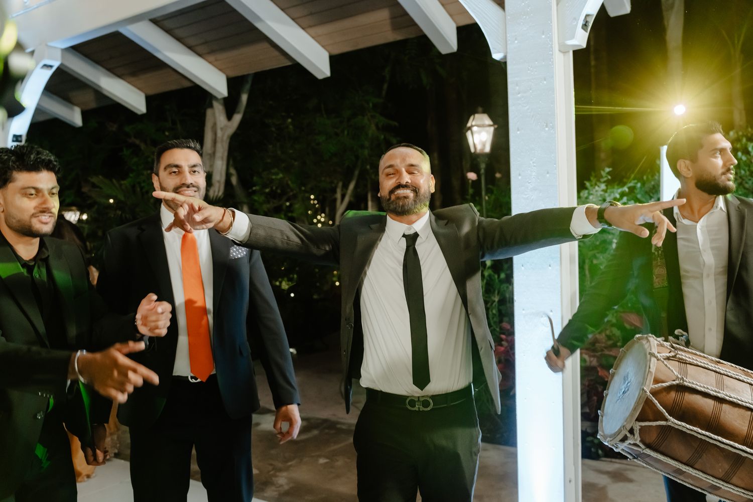 Groomsmen in dark suits pose together under a white pergola during evening celebrations.