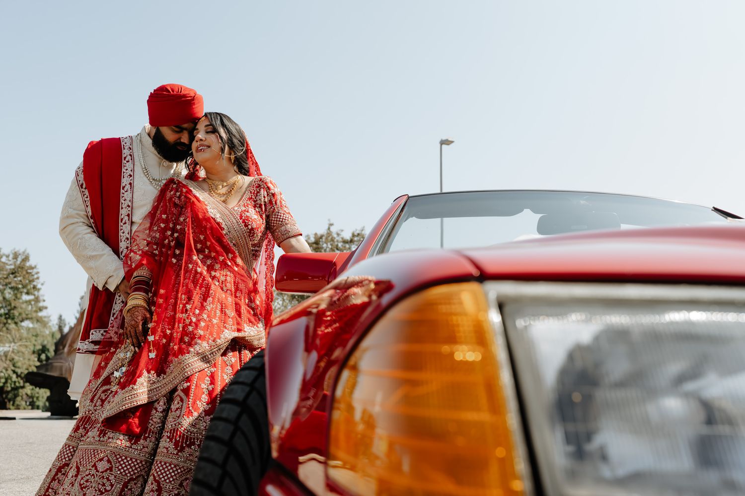 Traditional Indian wedding couple standing beside a classic red car.