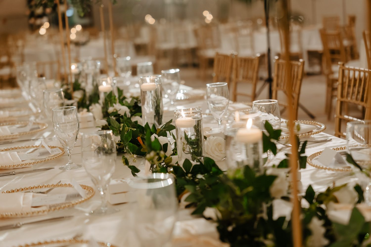 Elegant wedding reception tables decorated with white floral centerpieces, candles and greenery in a luxurious venue.