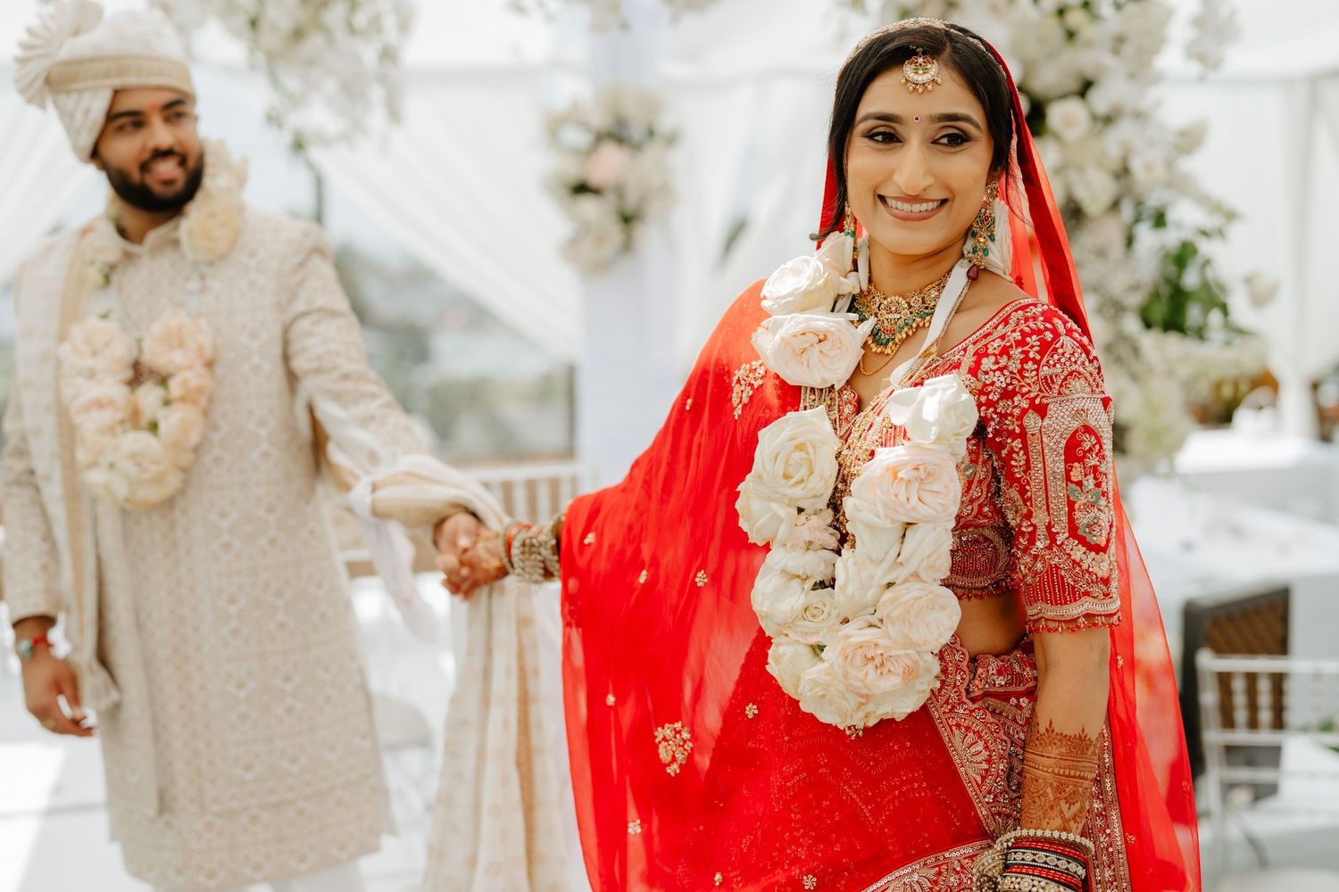 Indian bride in red lehenga with floral garland smiles as groom in white sherwani holds her hand at wedding ceremony.Mitul and Shivani at th