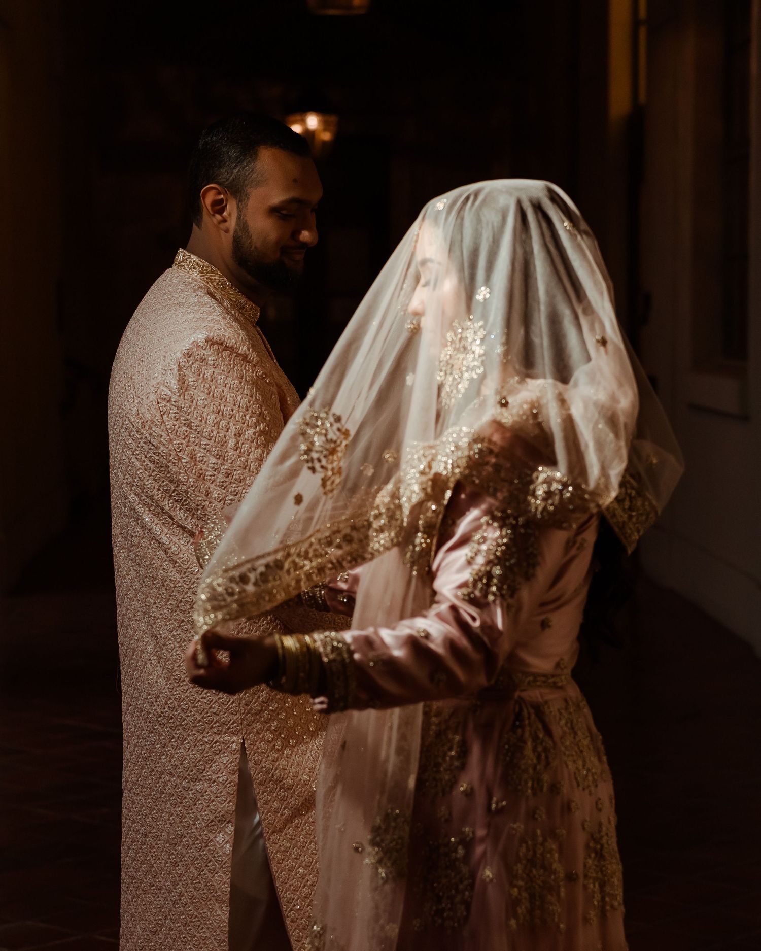 A couple in traditional South Asian attire stands together in a dimly lit indoor setting.