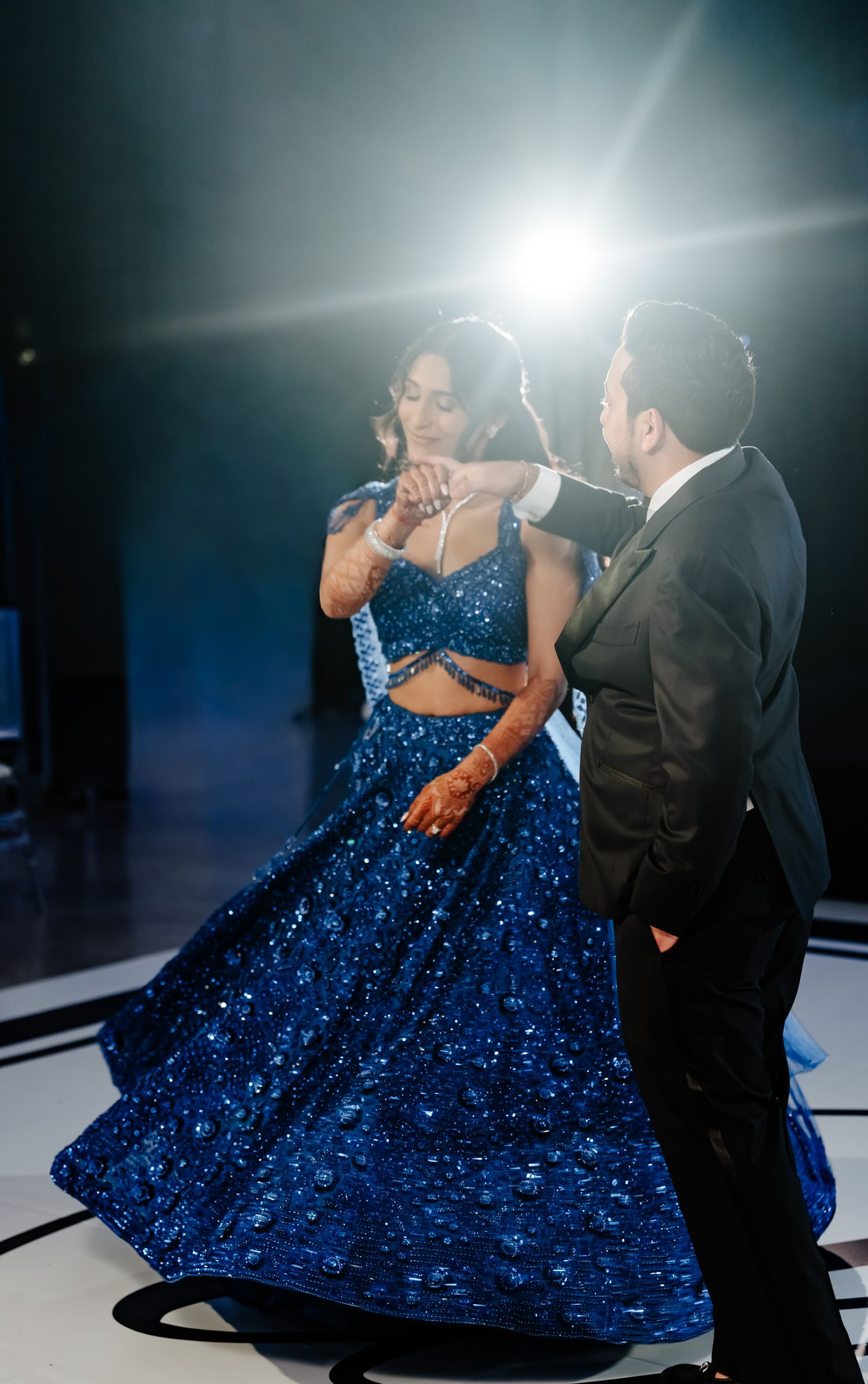 Couple dancing in elegant attire with woman wearing sparkling blue gown under dramatic lighting.
