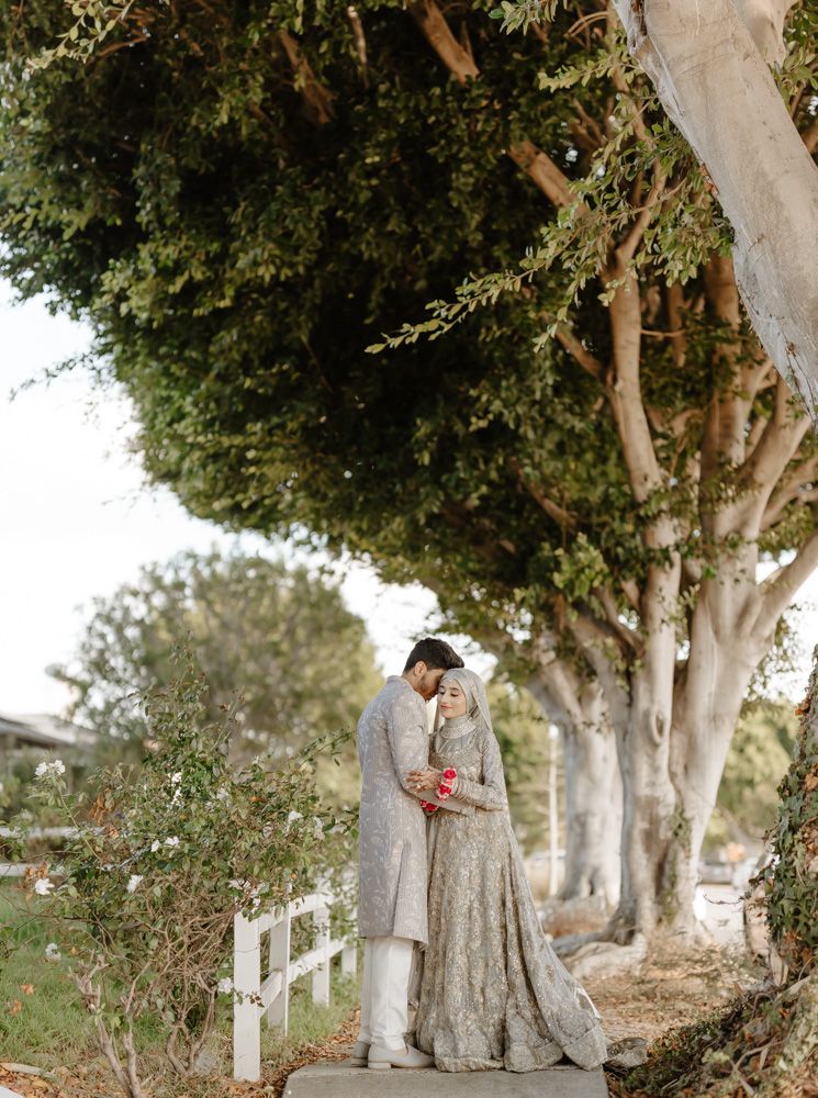 South Asian couple in matching silver embroidered traditional attire share a romantic moment under lush green trees outdoors.
