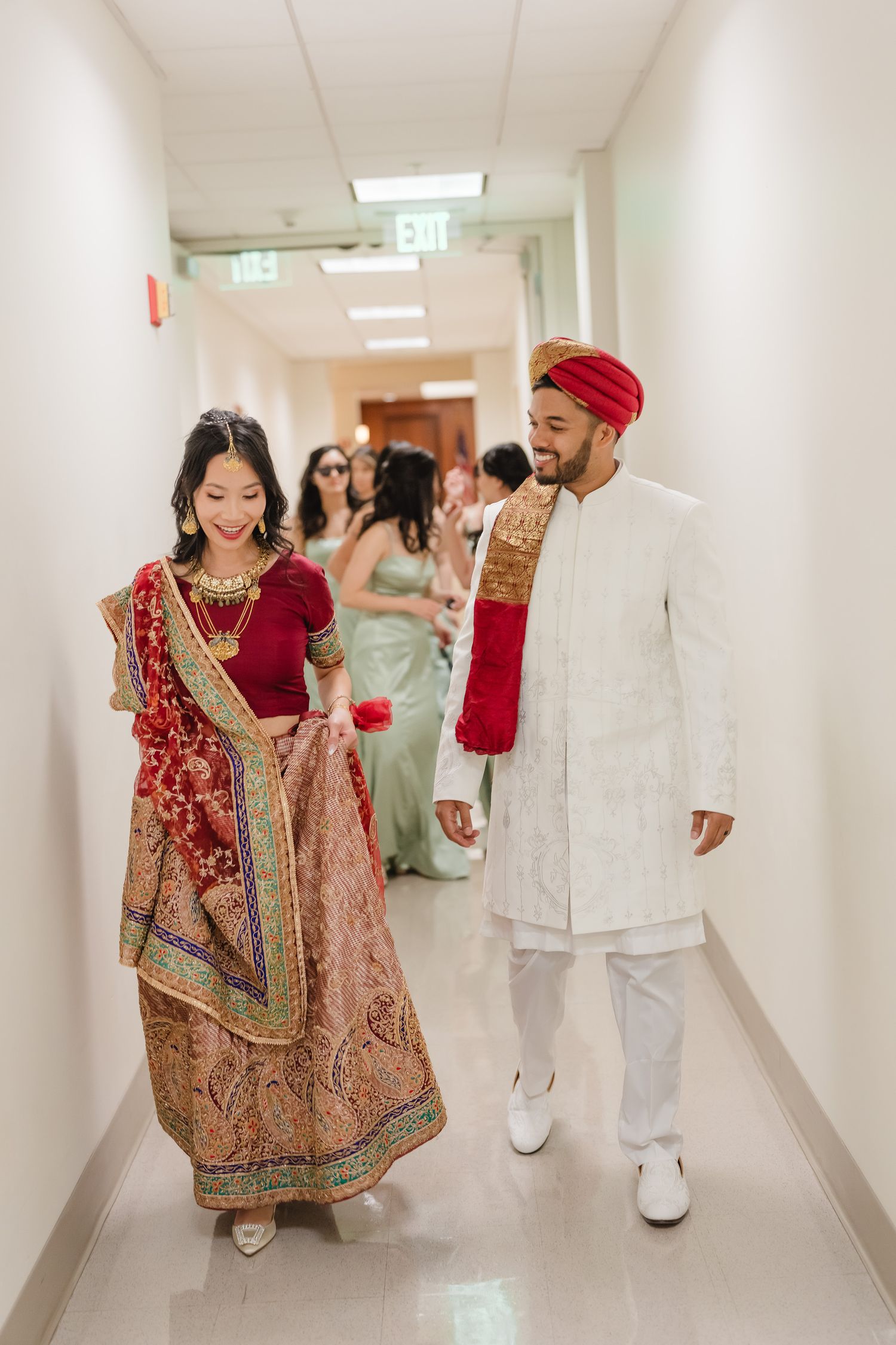 A couple in traditional Indian wedding attire walking down a hallway with guests in the background.