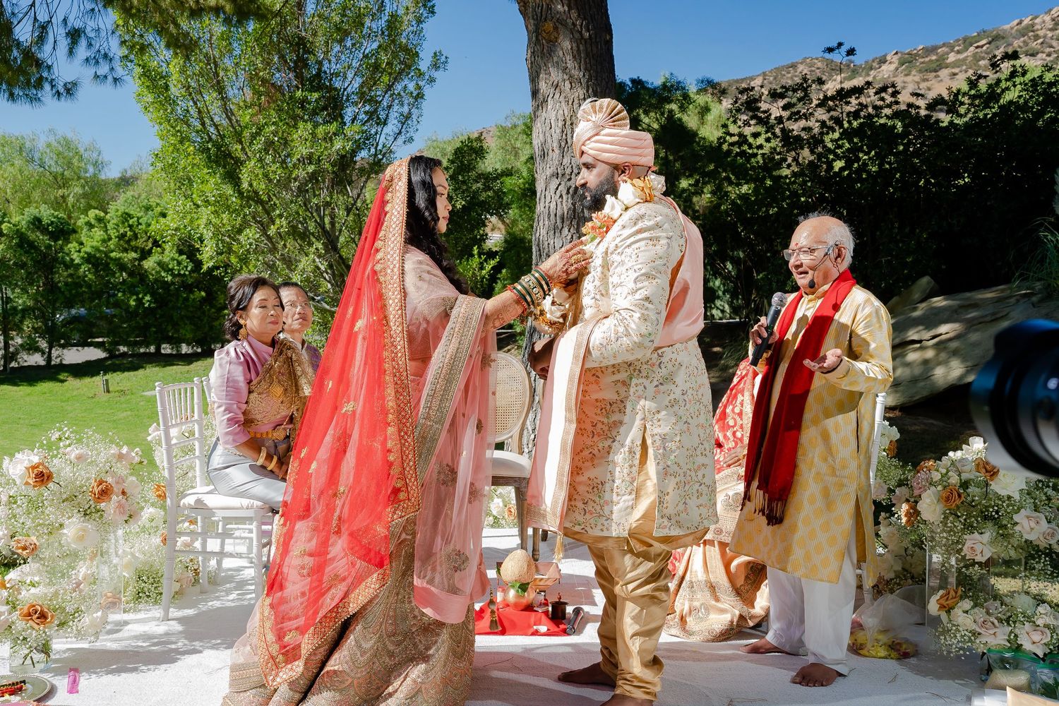 Traditional Indian wedding ceremony takes place outdoors in a garden setting with vibrant red and gold decor.