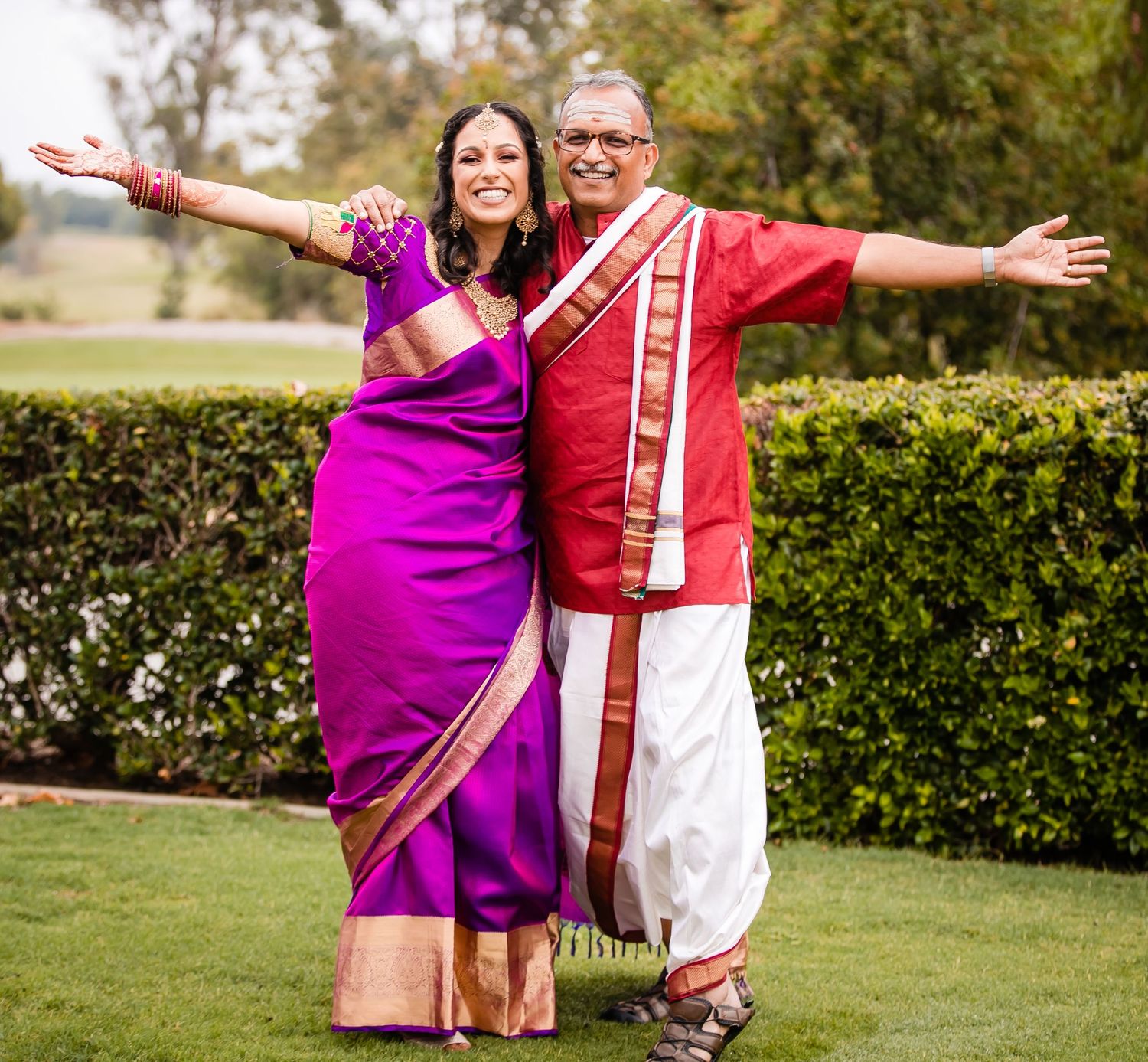 A couple in traditional Indian attire strikes joyful poses with outstretched arms in a garden setting.