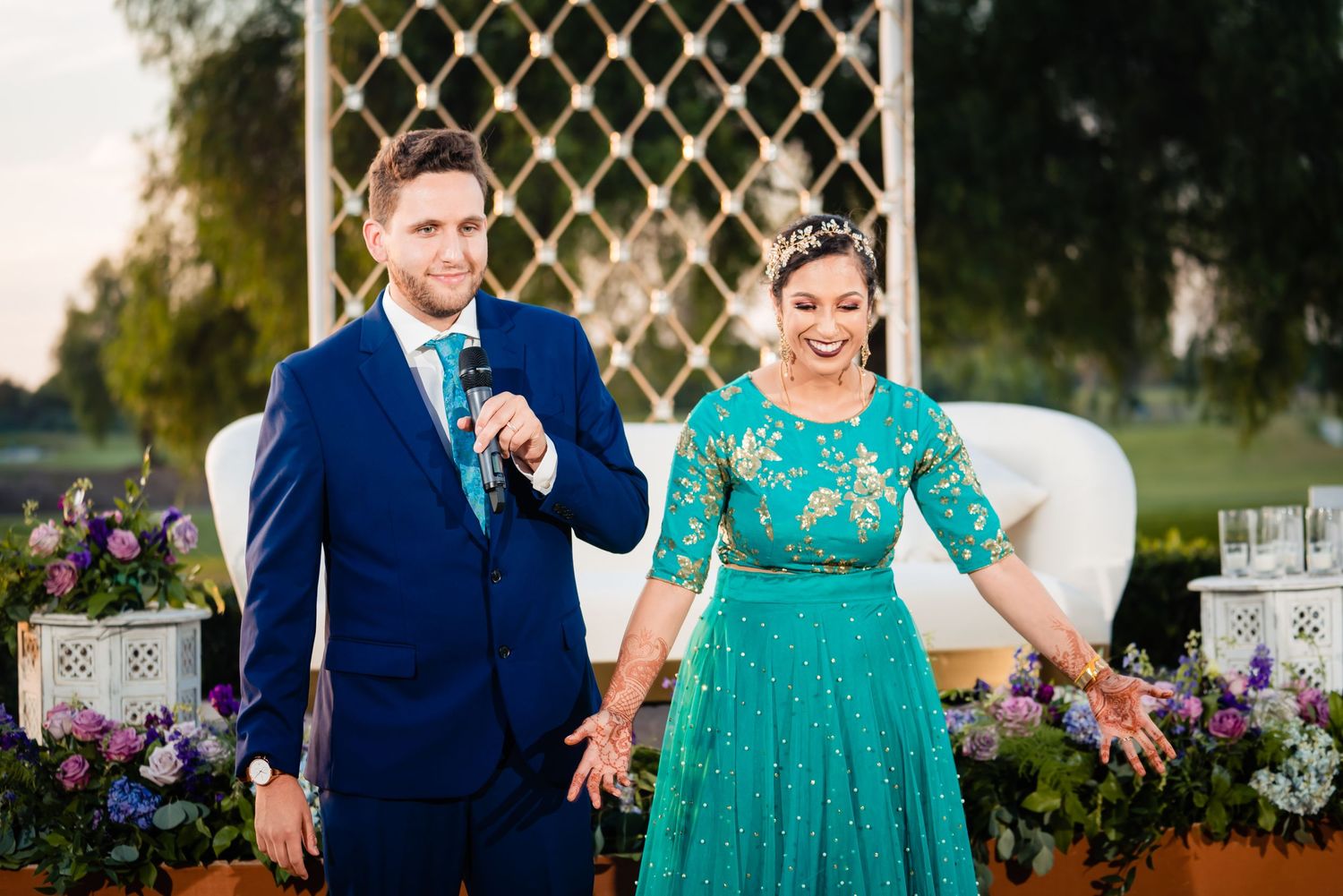 A couple in navy blue and emerald green formal attire pose together at an outdoor wedding venue with floral decorations.