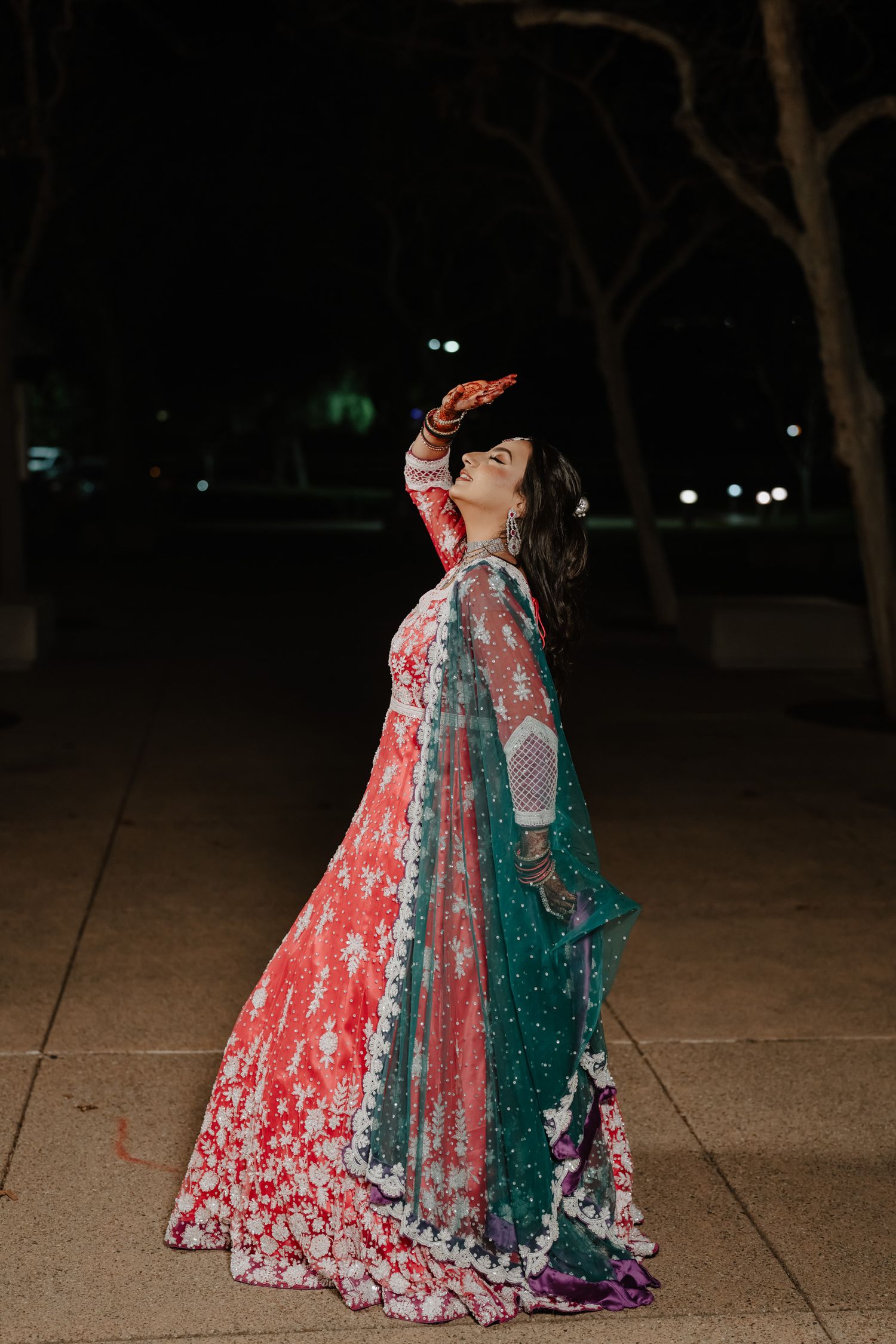 A person in an ornate red and green traditional Indian lehenga poses dramatically at night on a sidewalk.