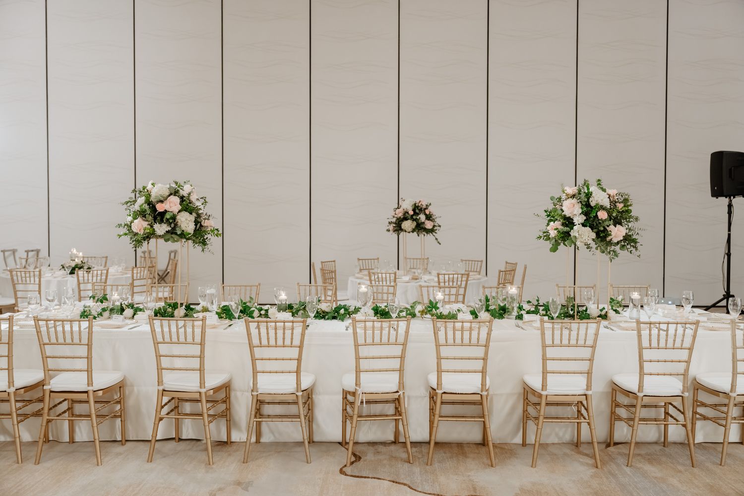 Elegant wedding reception table with gold chiavari chairs, white linens and floral centerpieces against a neutral wall.